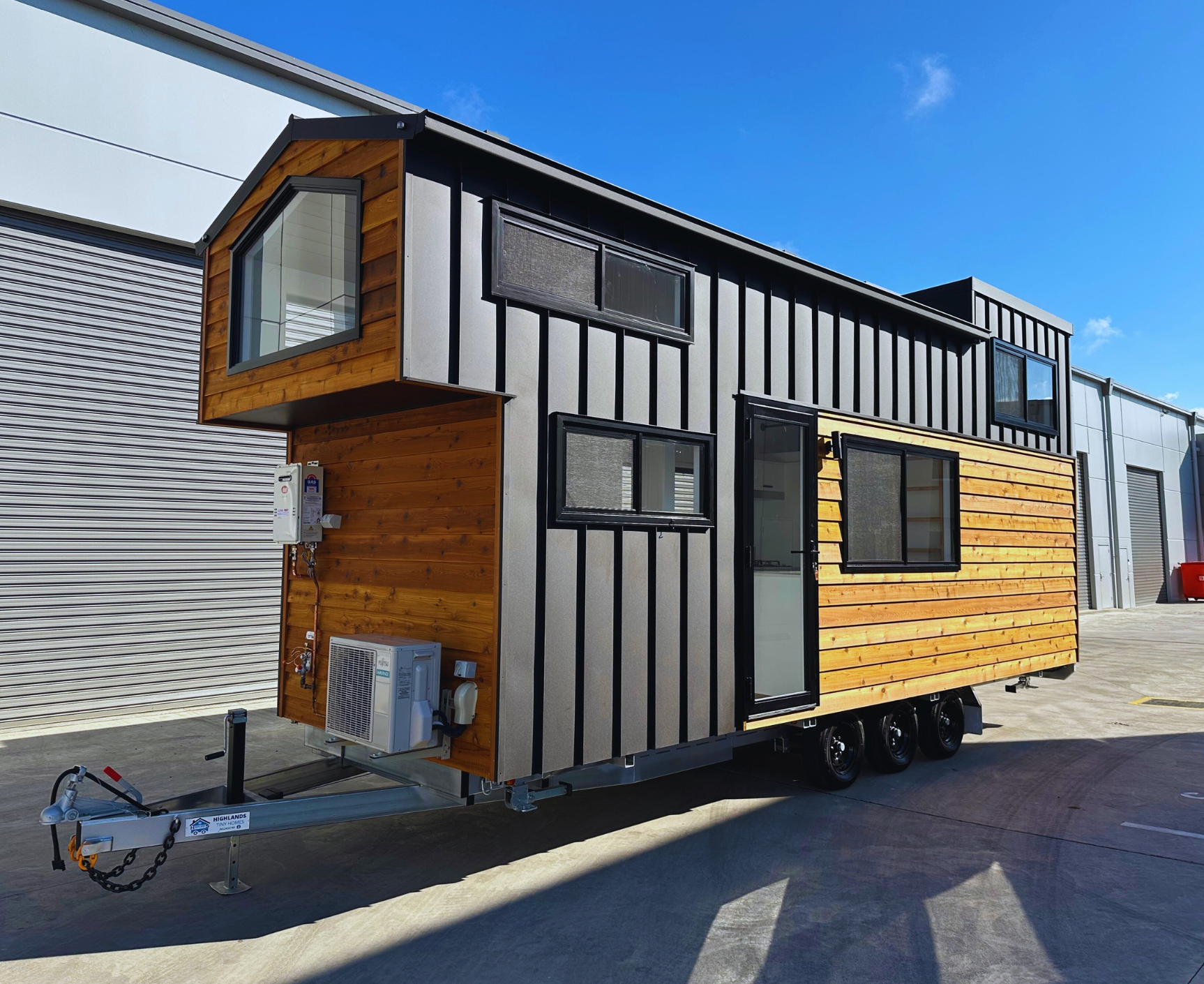 Tiny wooden house on wheels with open door, on green grass with a cloudy blue sky.