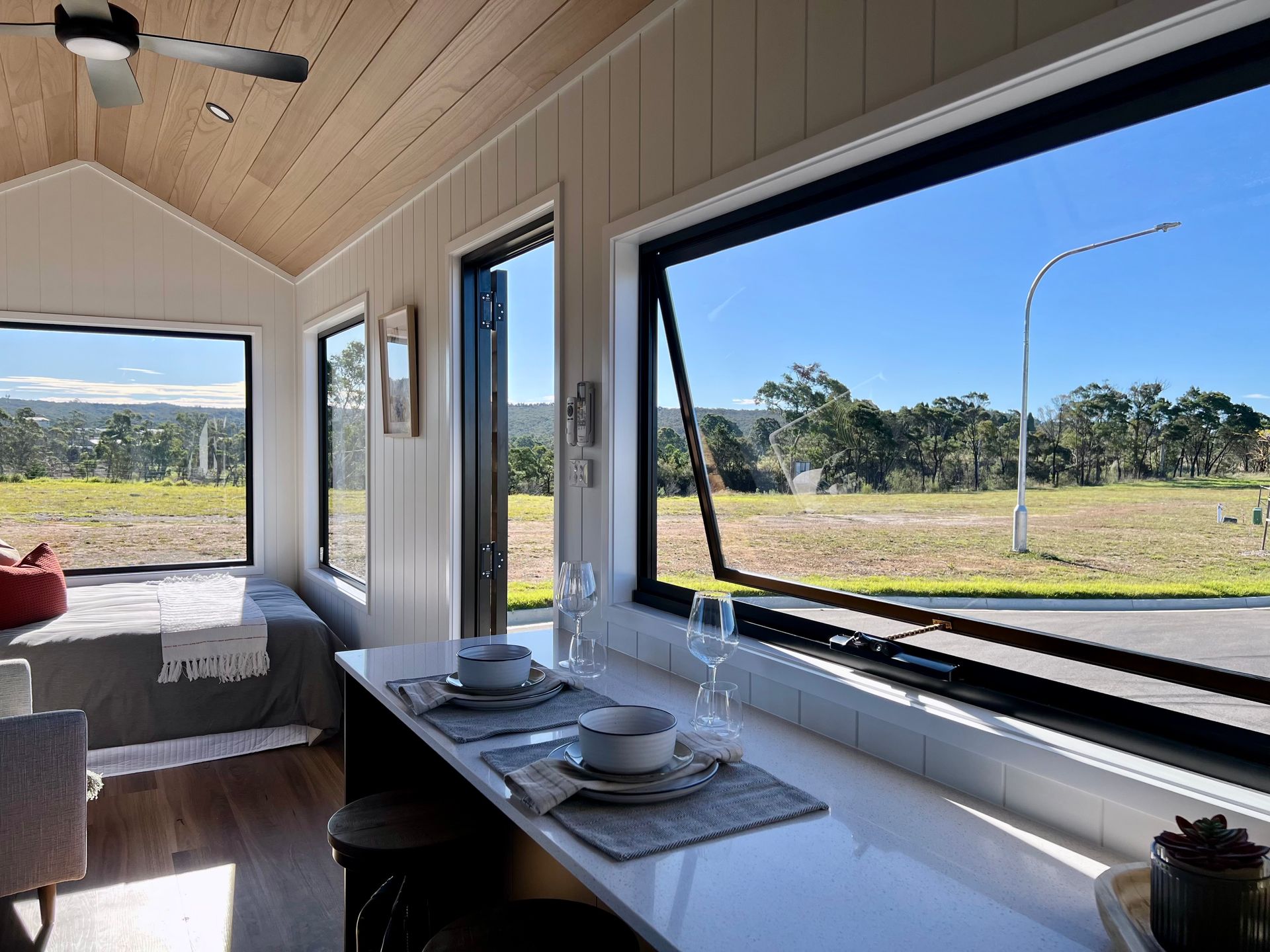 Interior of a tiny house with large windows overlooking a grassy field and blue sky. White walls and counter.
