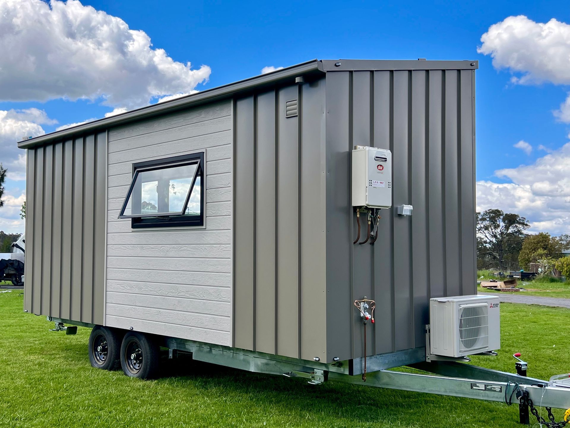 Tiny, modern house on wheels, gray and white exterior, parked on grass, sunny day.