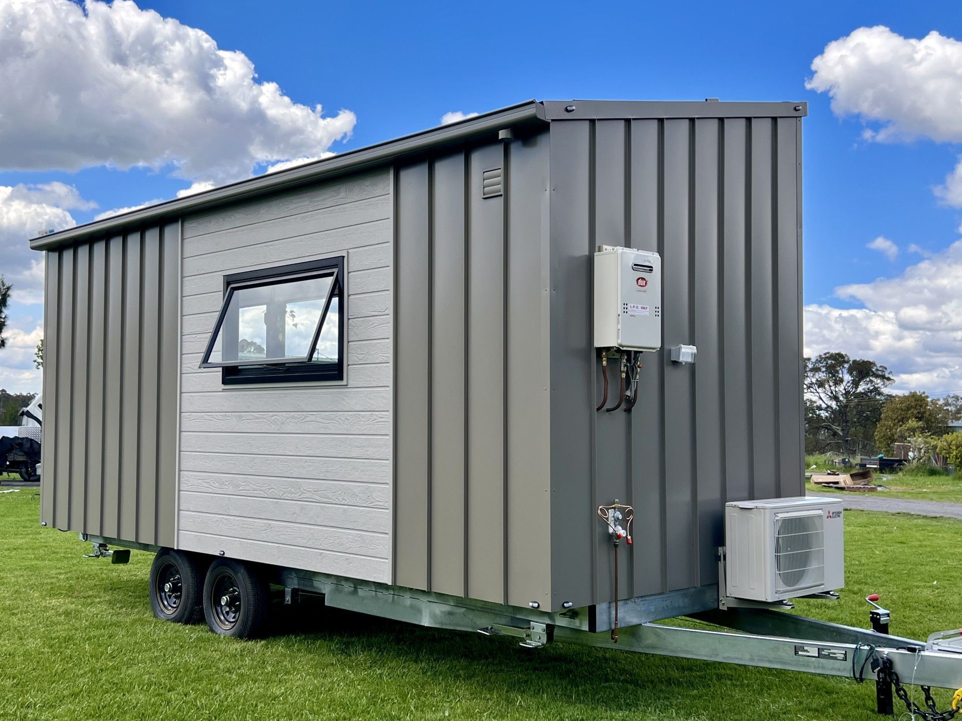 Tiny house on wheels with gray and white siding, sitting on a grassy field, under a blue sky.