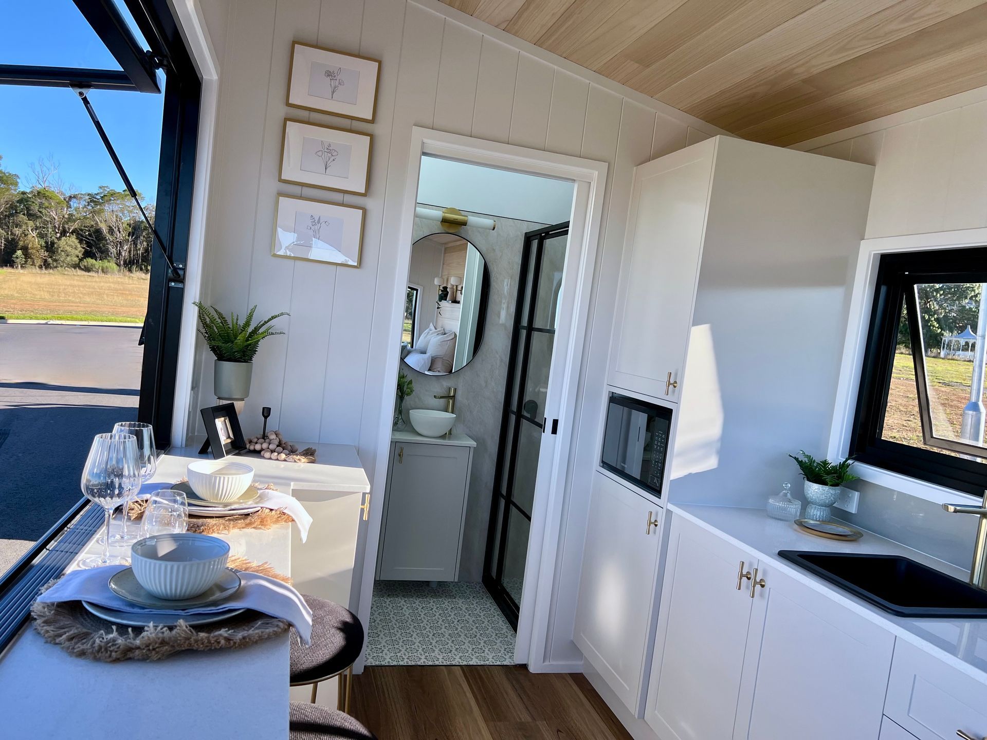 Interior of a tiny home with kitchen, dining area, and a bathroom visible. White walls, wooden floors, and natural light.