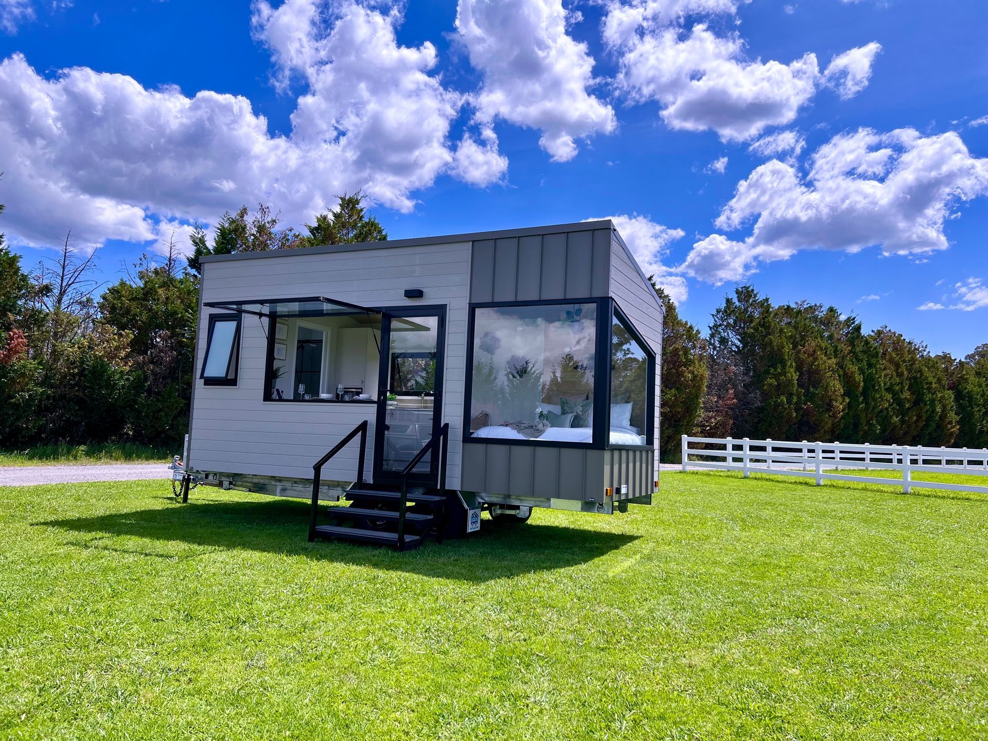 Tiny house on wheels, gray and white exterior with large windows, set in a grassy field under a blue sky.