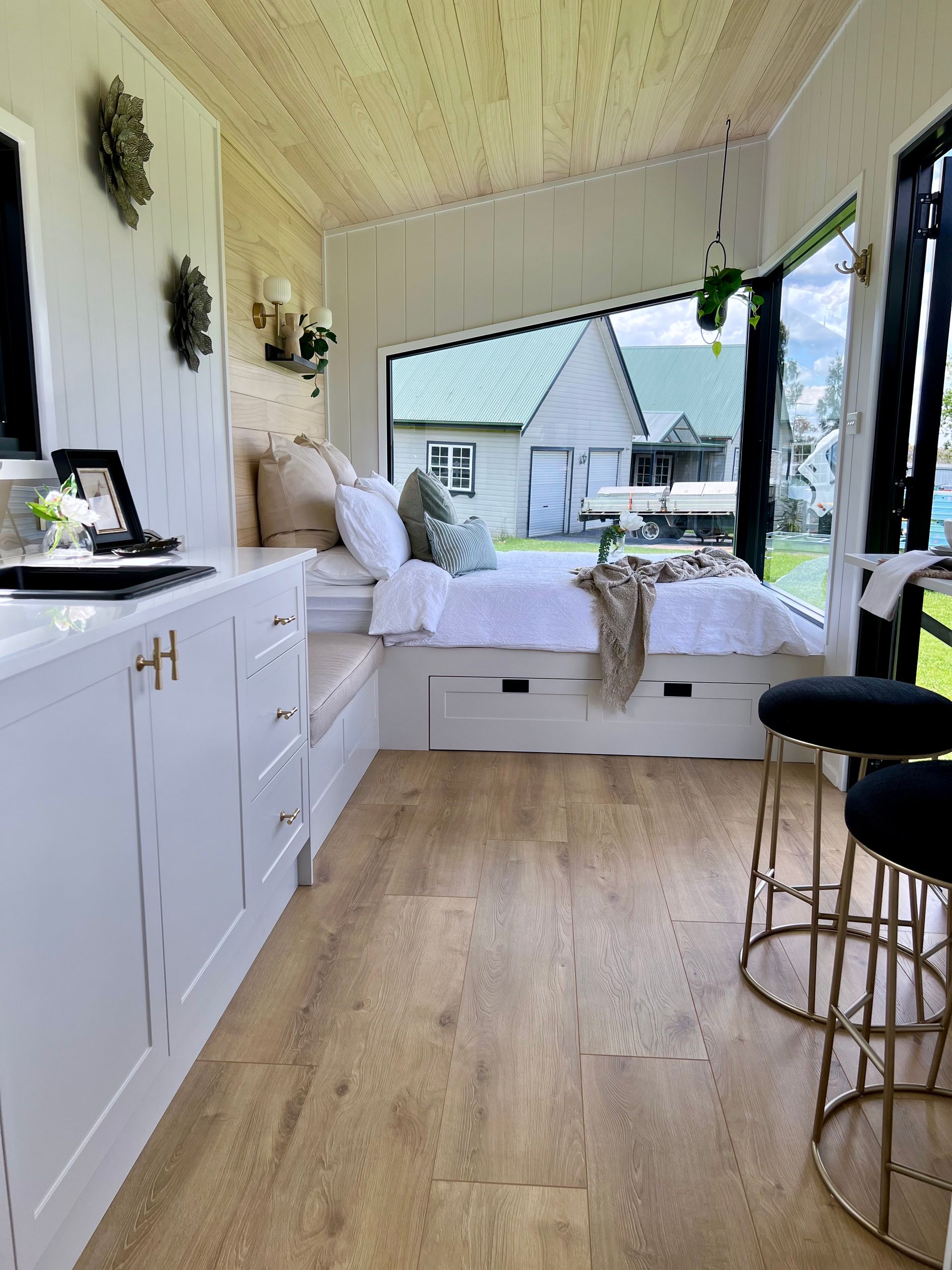 Tiny home interior with a bed nook, white cabinetry, and large window overlooking a green landscape.