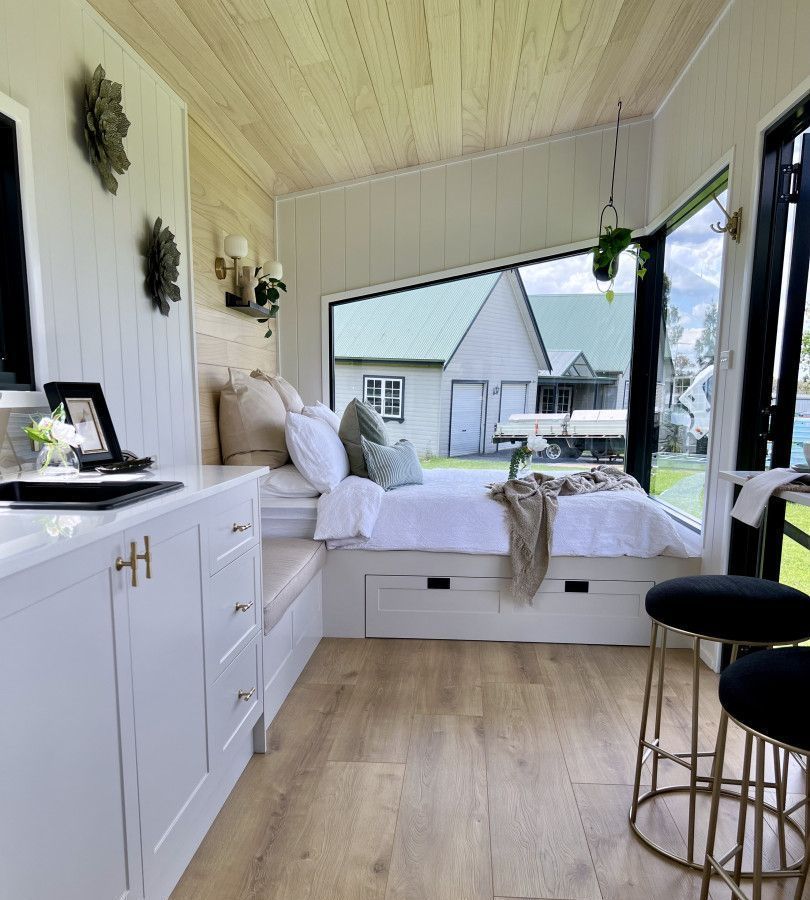 Tiny house on a trailer, with a wooden deck and black siding, parked in grassy area.