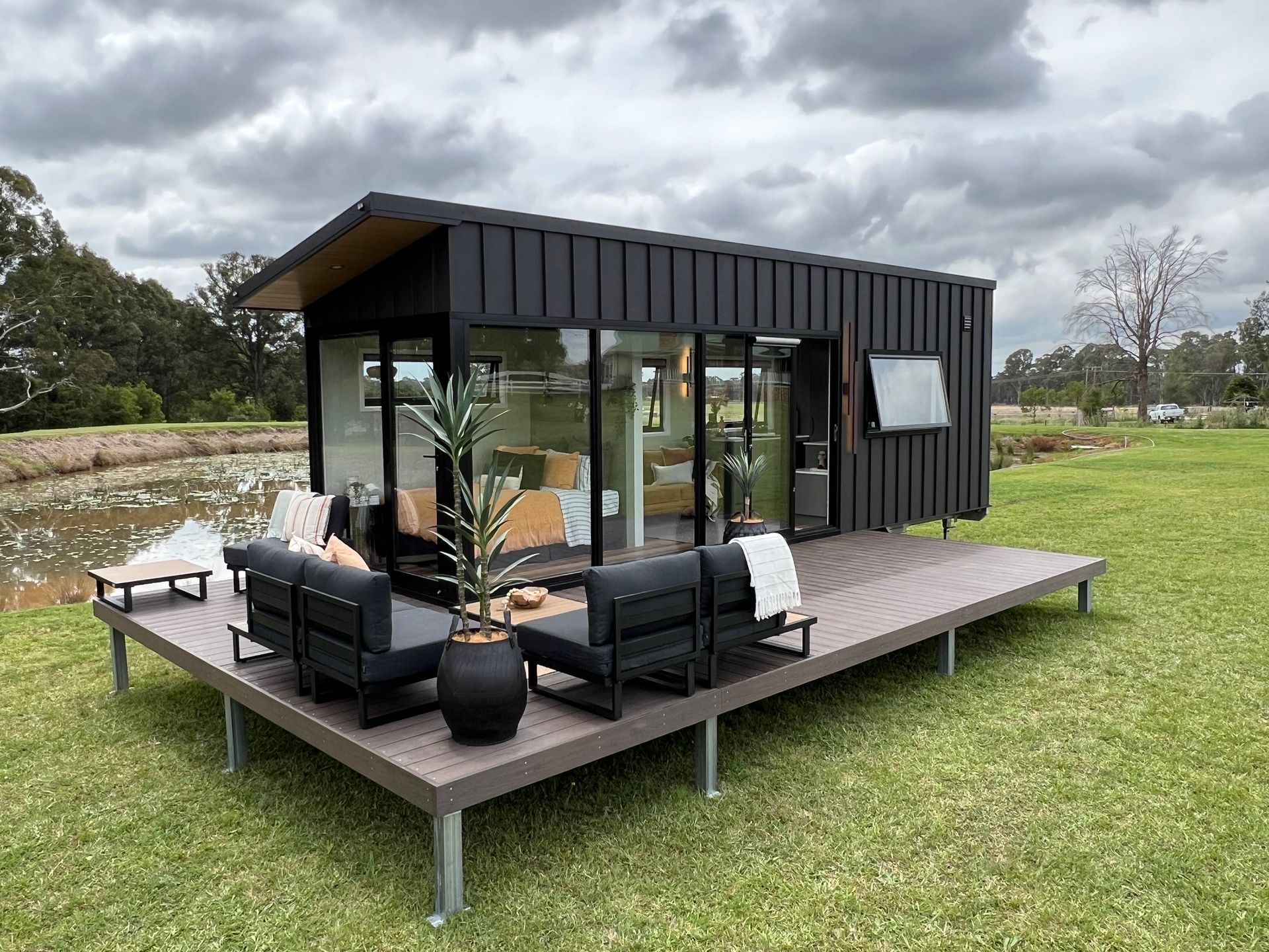 Tiny modern house with deck, black exterior, large windows, and outdoor seating on a grassy area next to a pond.