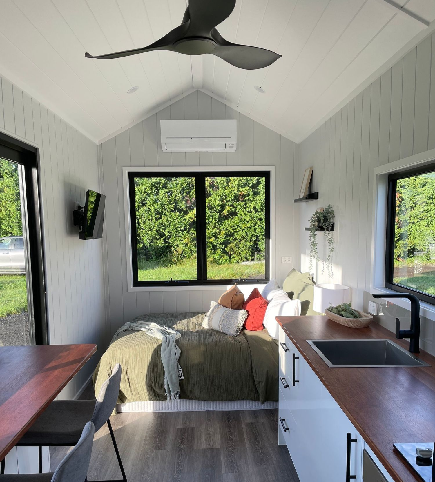 Cozy tiny home interior with bed, kitchen, windows, and dark wood countertops.