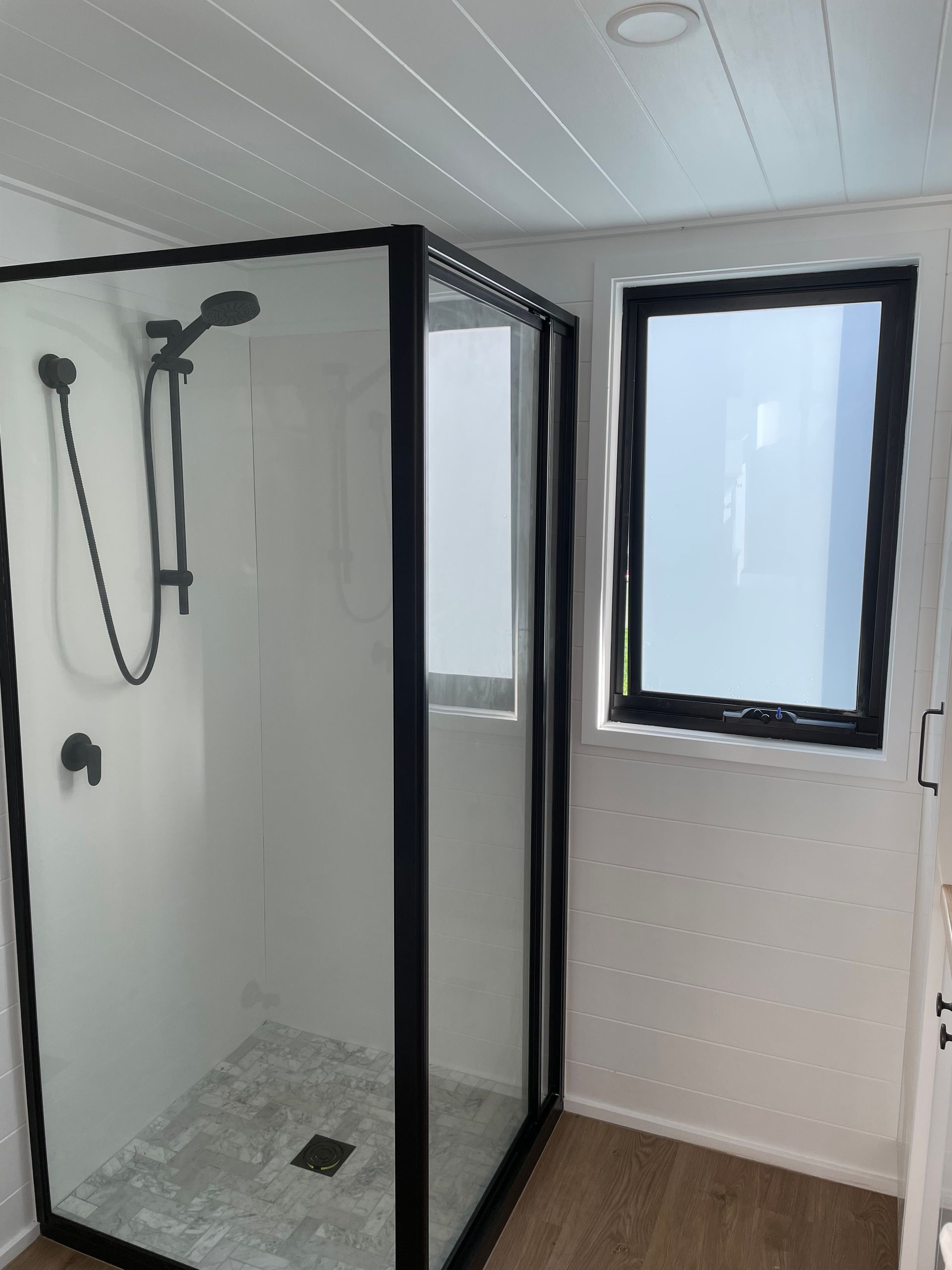 Shower with black frame and fixtures next to a window in a white bathroom.
