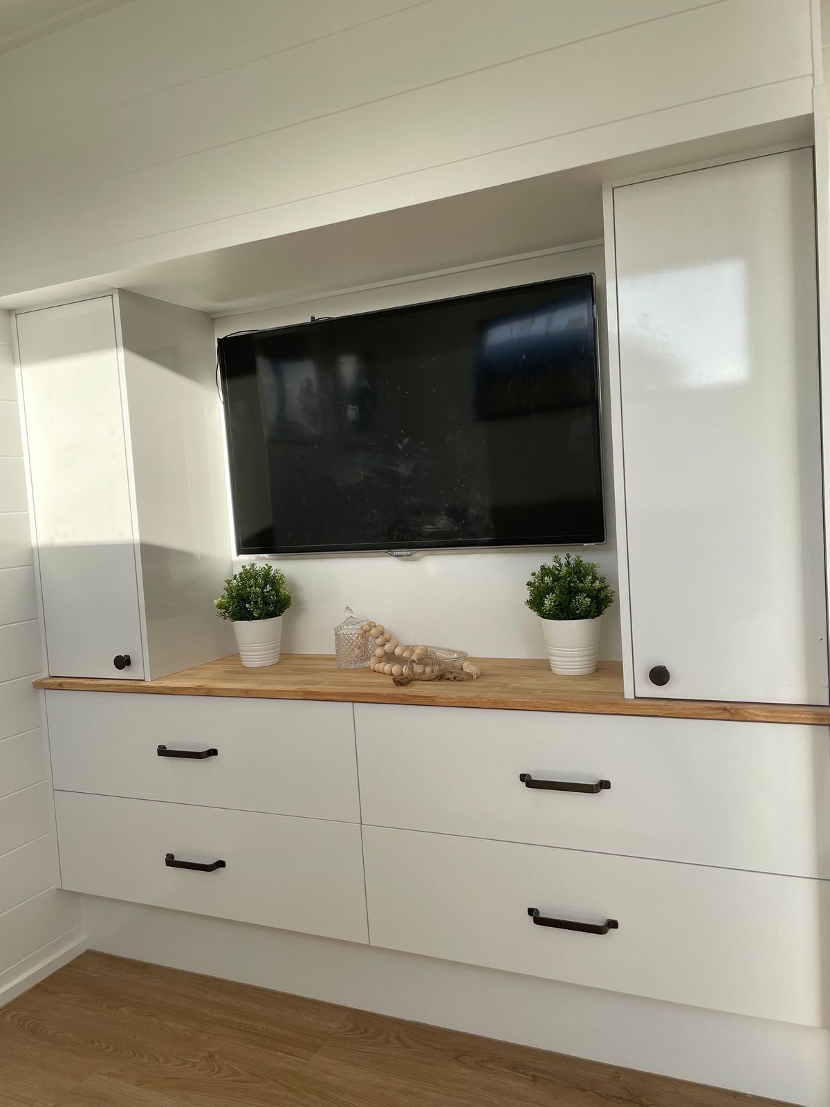 White built-in entertainment center with drawers, cabinets, TV, and plants on a wooden surface.