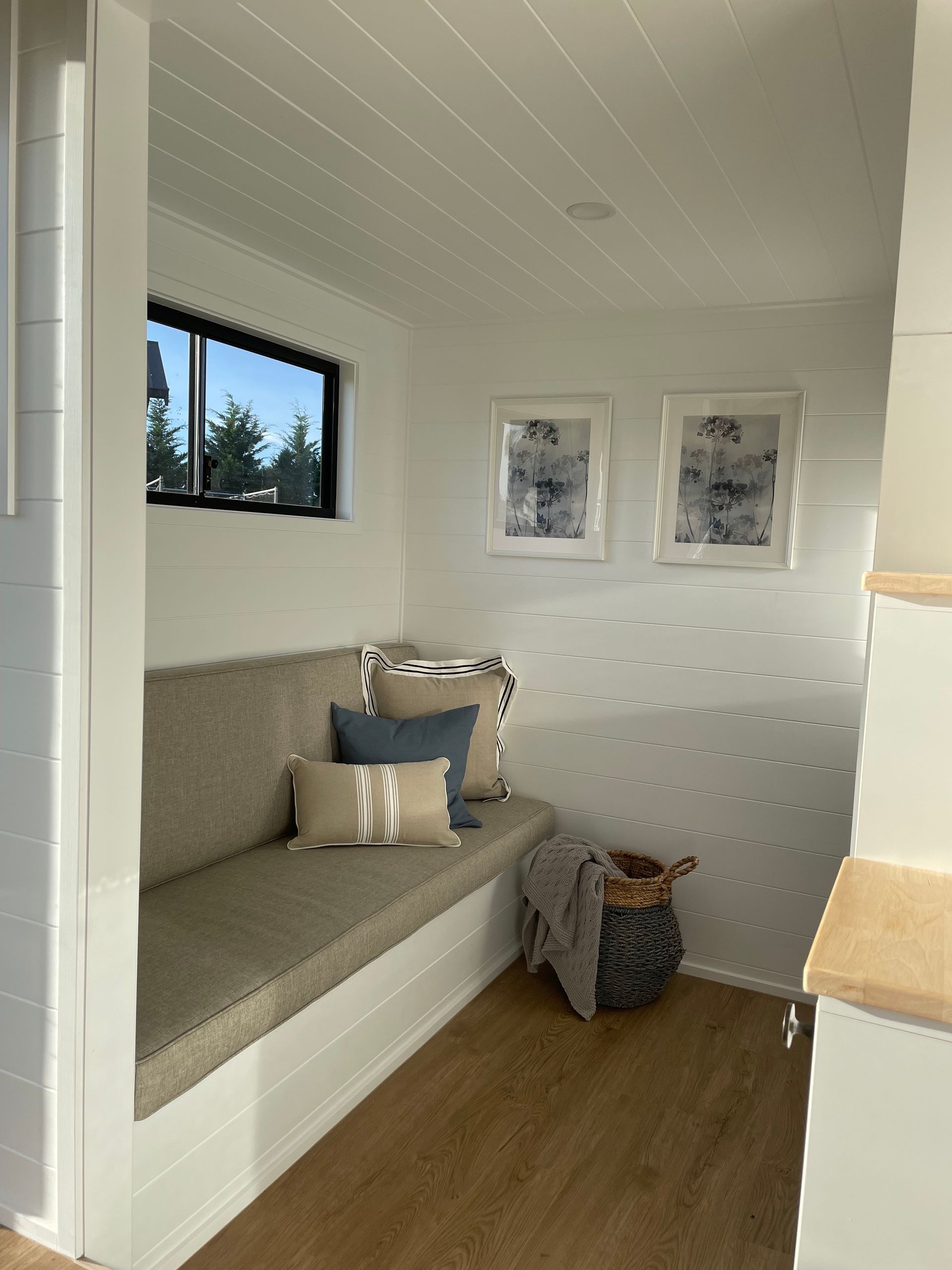 Cozy, white-walled nook with a bench, window, and artwork. A basket with blanket sits on wooden floor.