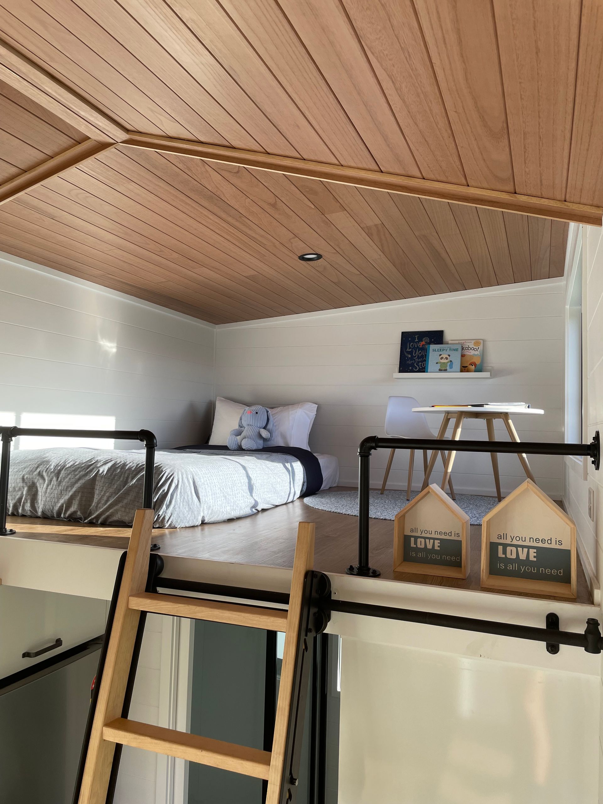 Loft bedroom with a bed, desk, ladder, and a wooden ceiling.