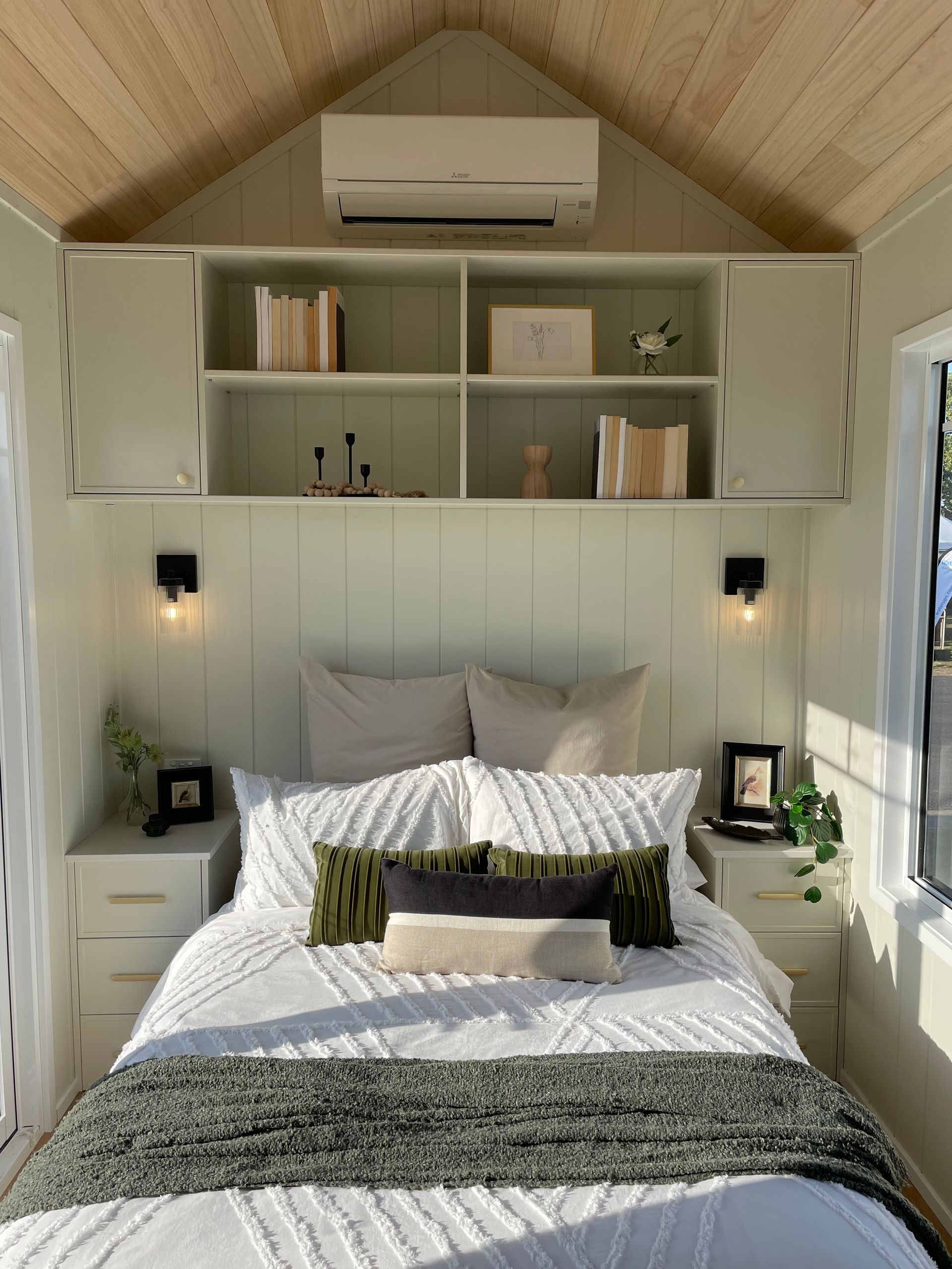 Cozy bedroom with white bed, green accents, storage cabinets, sconces, and a window, all in a light, airy space.