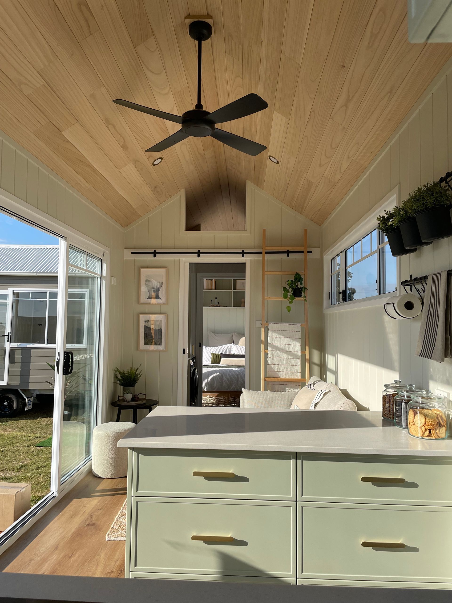 Interior of a tiny house with a light green kitchen, wood ceiling, and a view of the bedroom.