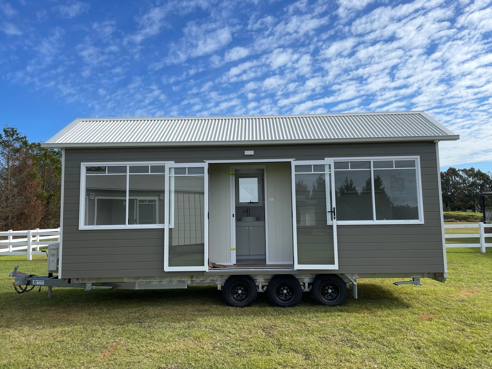 Gray tiny house on wheels with open doors, set on grass under a blue sky.