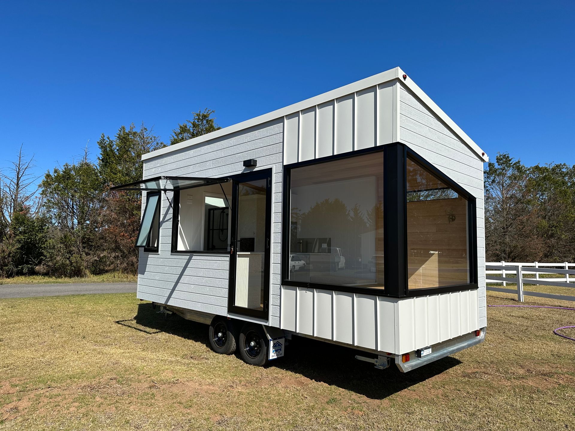 Tiny, light gray house on wheels with black framed windows, door, and a white fence behind it, outdoors on a sunny day.
