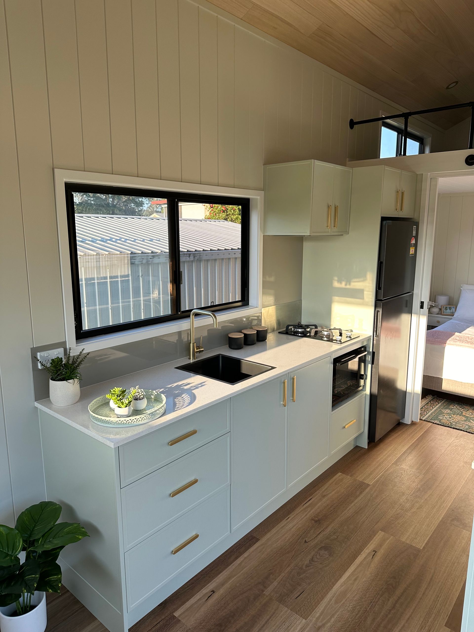 Tiny kitchen with light green cabinets, gold hardware, and a black sink. A window overlooks a backyard.
