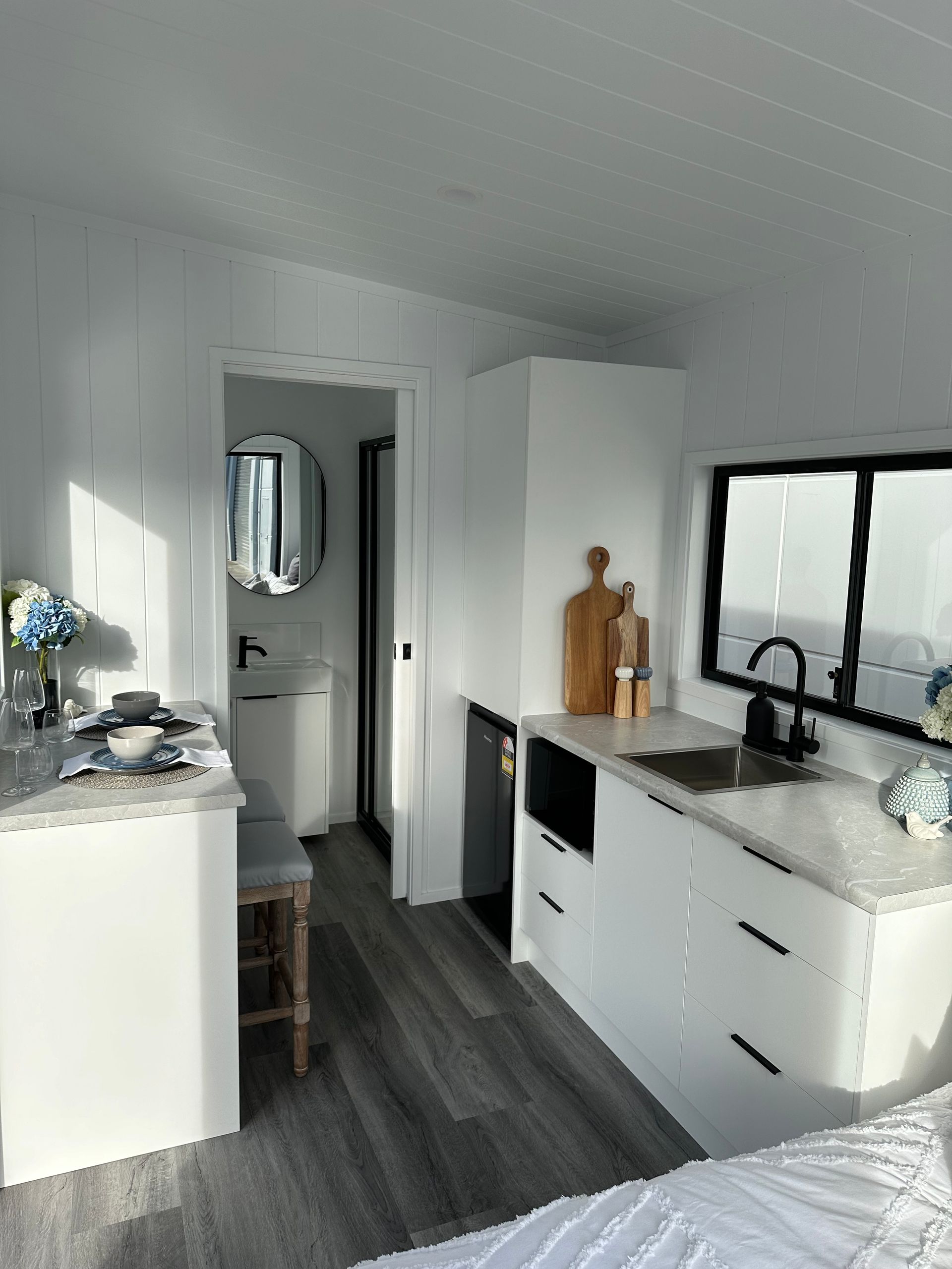 Interior of a tiny, modern, white kitchen and bathroom. Features a sink, cabinets, and a small round mirror.