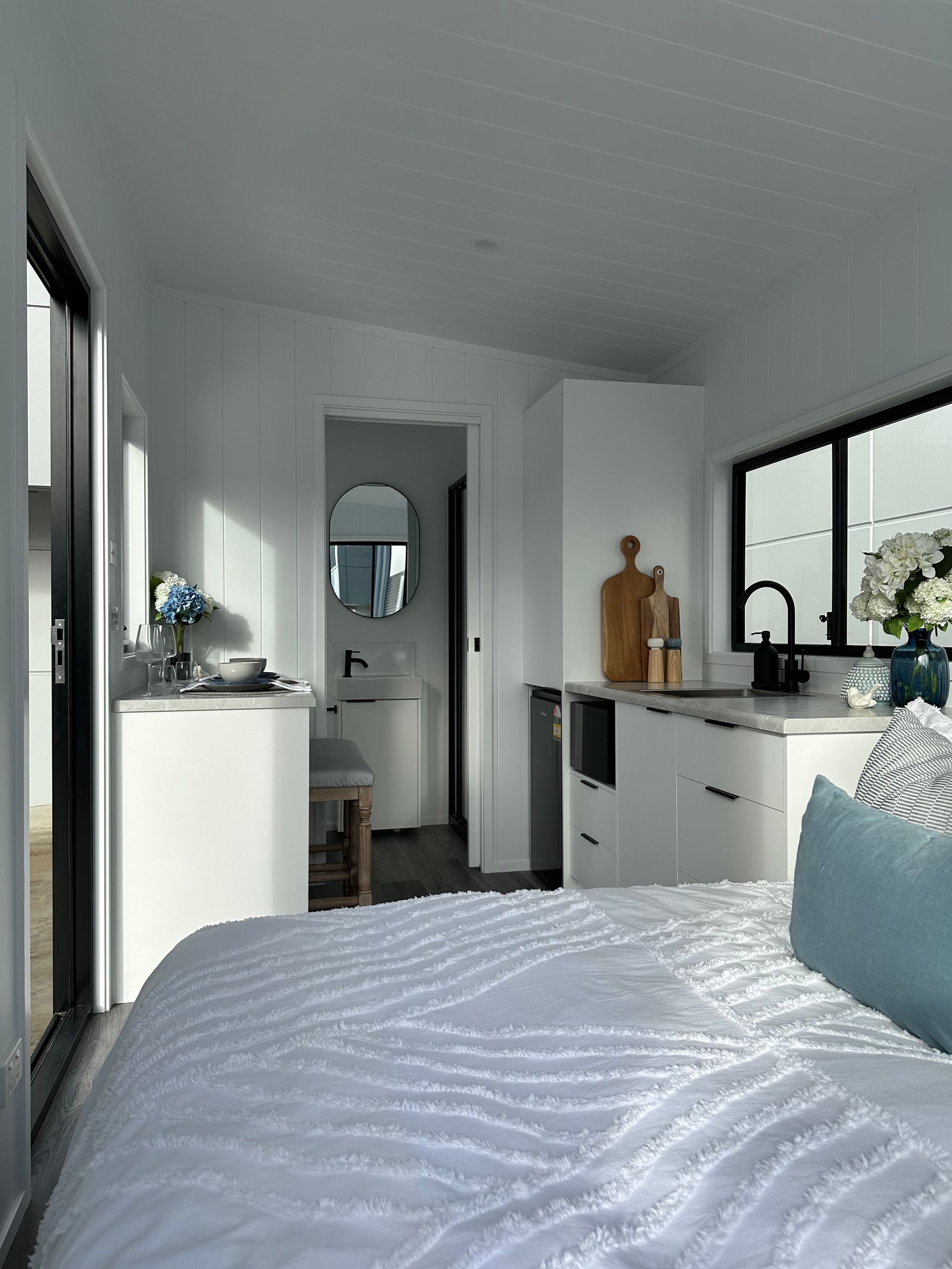 Cozy white tiny home interior with bed, kitchen, and bathroom; sunlit.