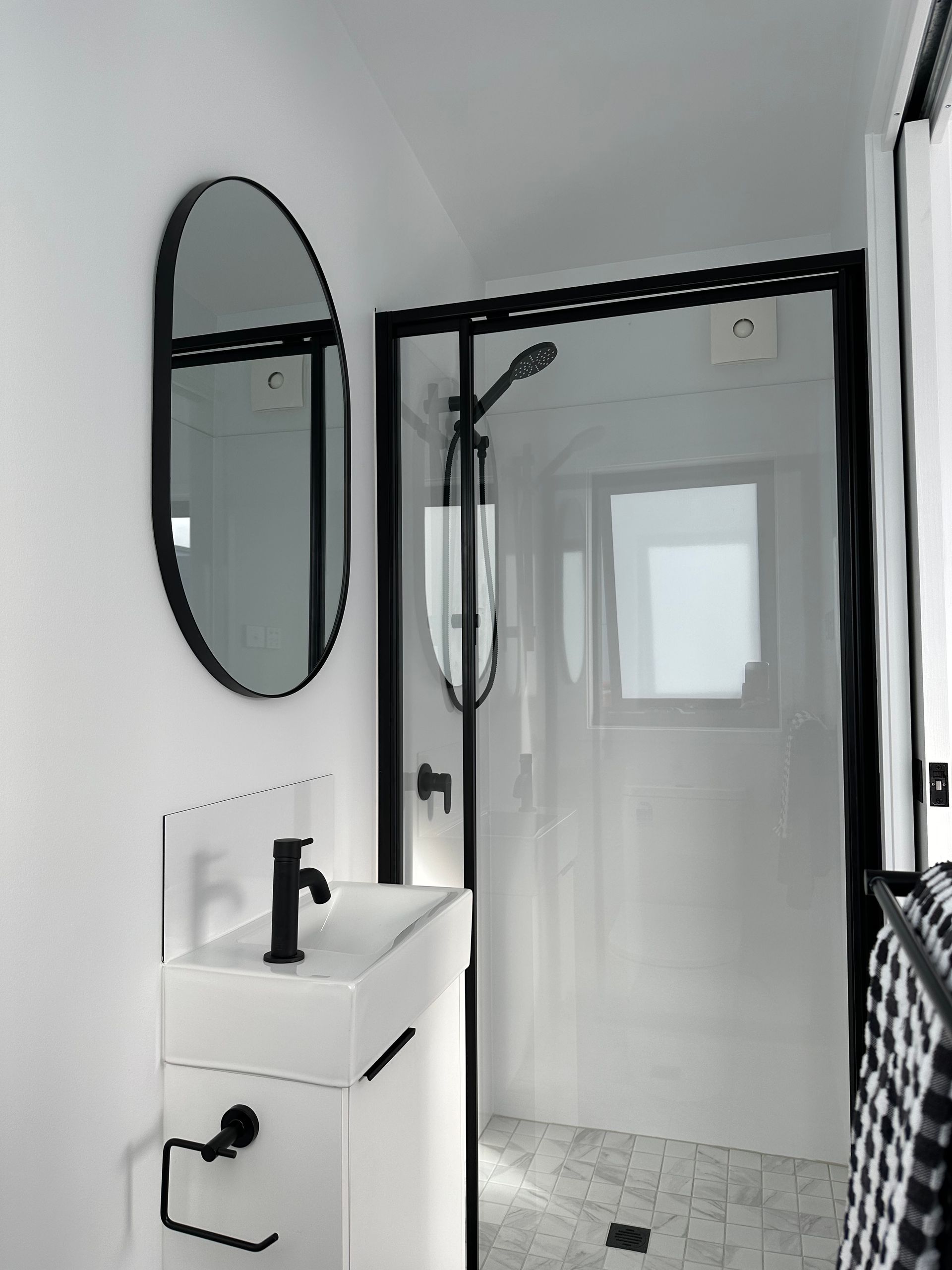 White bathroom with black fixtures: sink, mirror, shower. Black and white towel hanging.