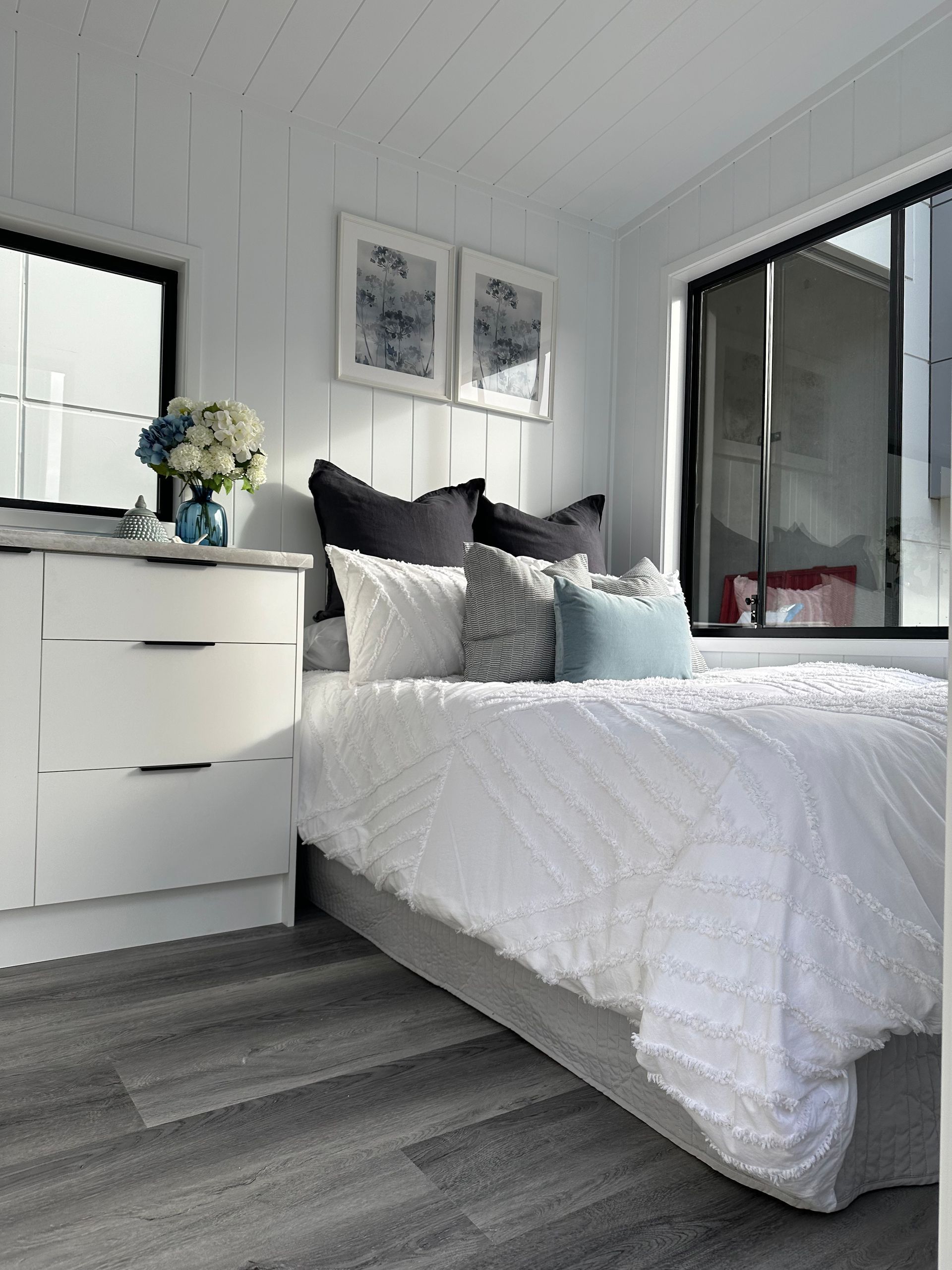 Bedroom with white walls, gray floor, bed, dresser, artwork, and large window.