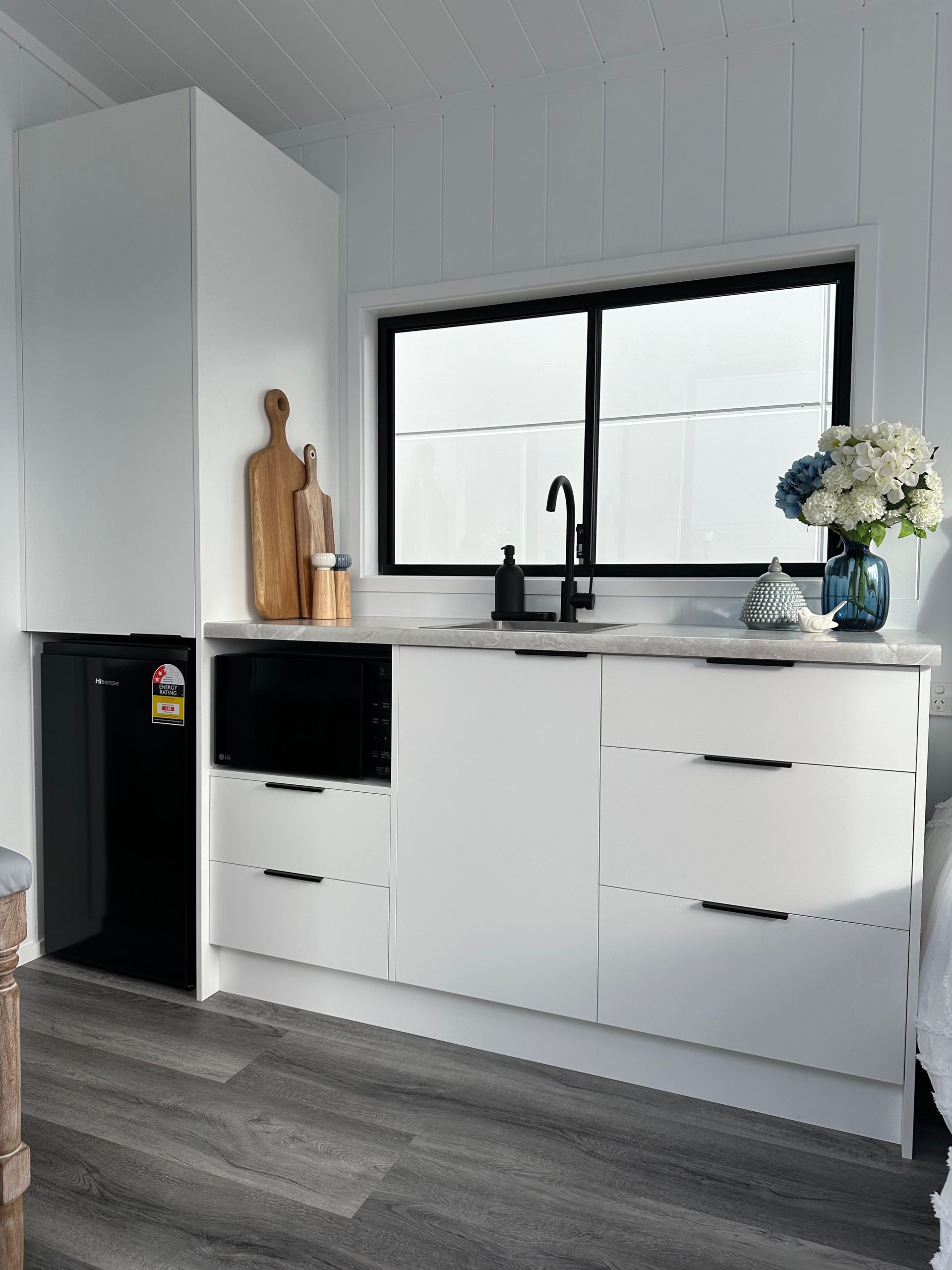 A small, white kitchenette with a black window, sink, and mini-fridge. It has wooden cutting boards and flowers.