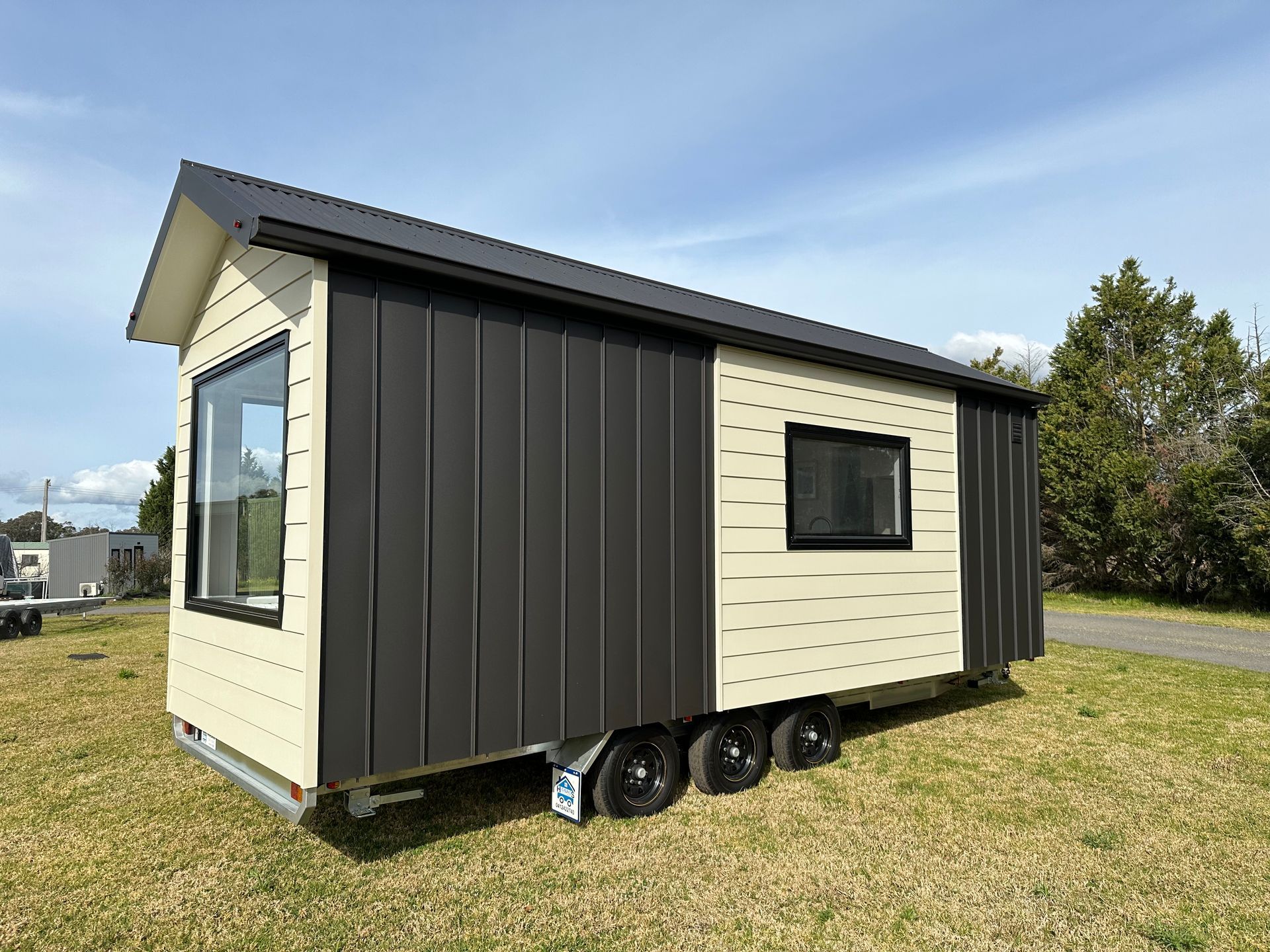 Tiny house on wheels with black and cream siding on a grassy area under a blue sky.
