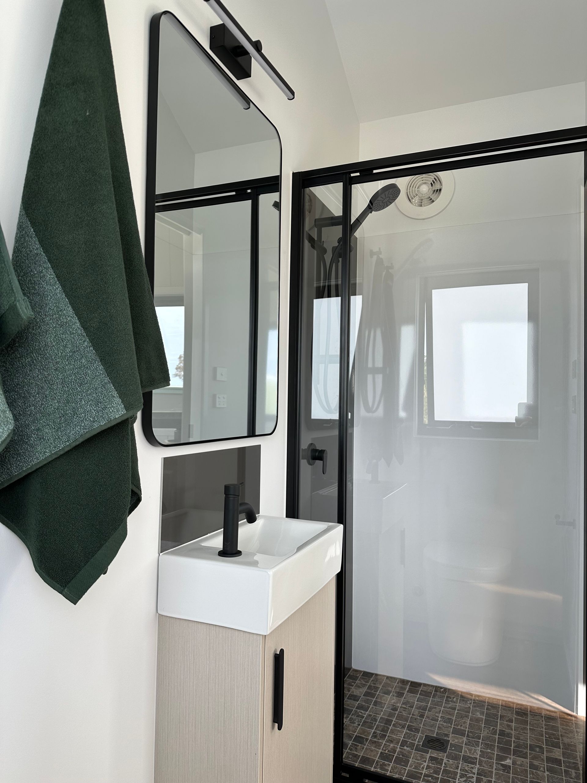 Small modern bathroom with a black-framed mirror, white sink, black faucet, and glass shower. Green towels hang on the wall.