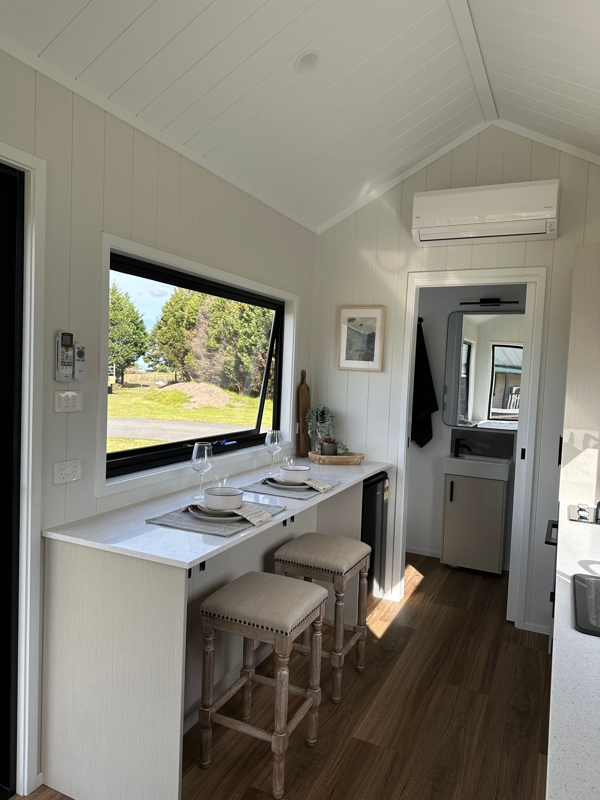 Interior of a tiny home with a bar counter, stools, and a window overlooking green trees.