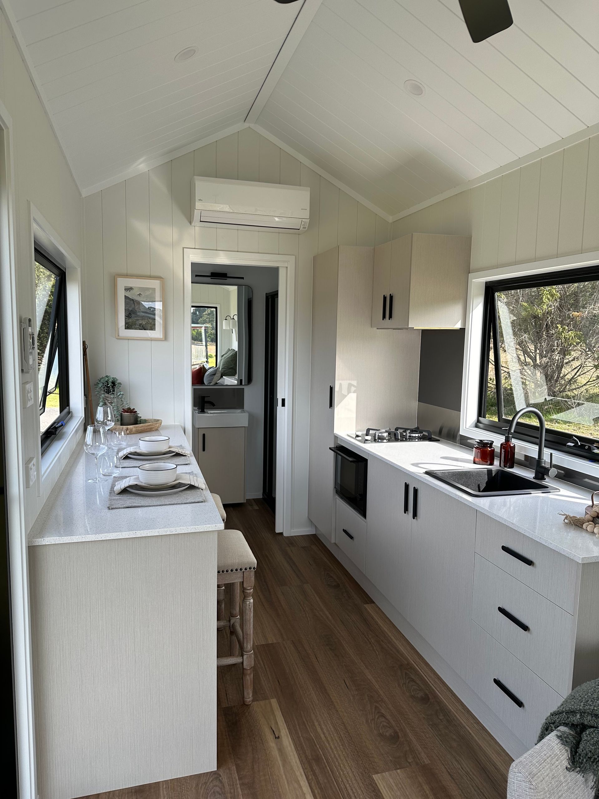 Interior view of a tiny home kitchen. White cabinetry, counters, and walls. Wooden floor. Sink and stovetop are visible.