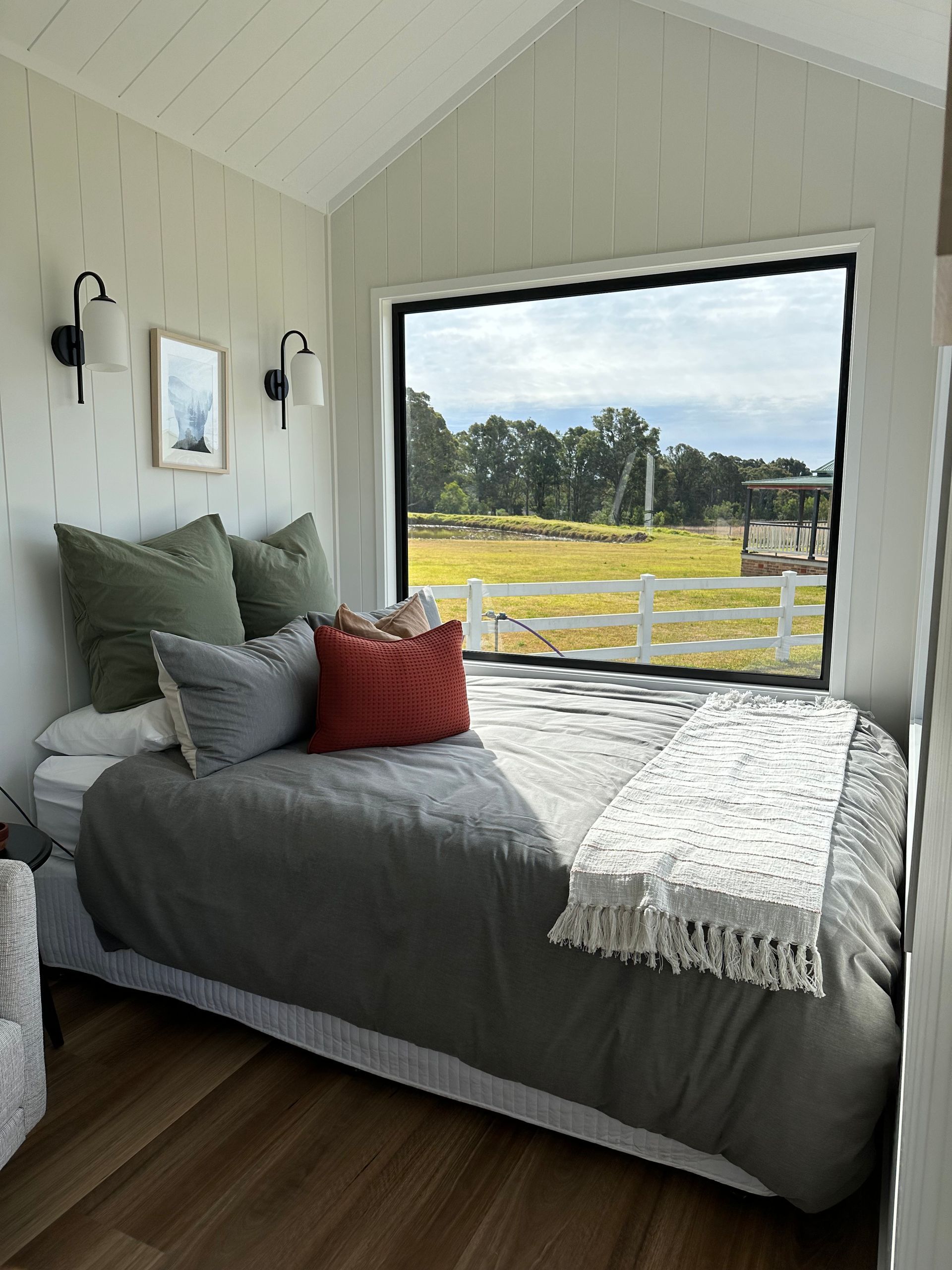 Bedroom with a bed, window with a view, green and red pillows, white walls, and black sconces.