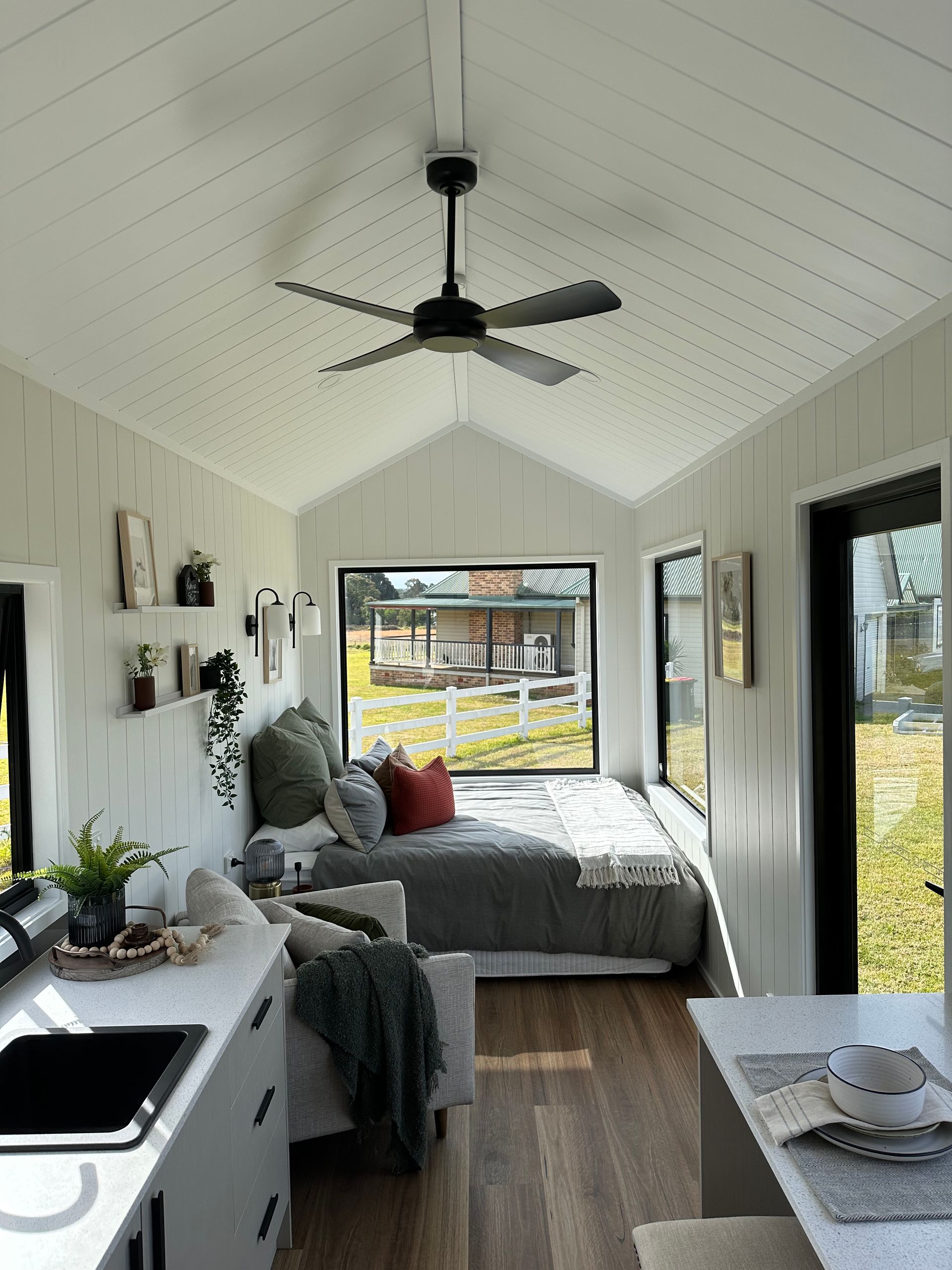 Small, bright interior of a tiny home with white walls, a bed, and a view outside through a window.