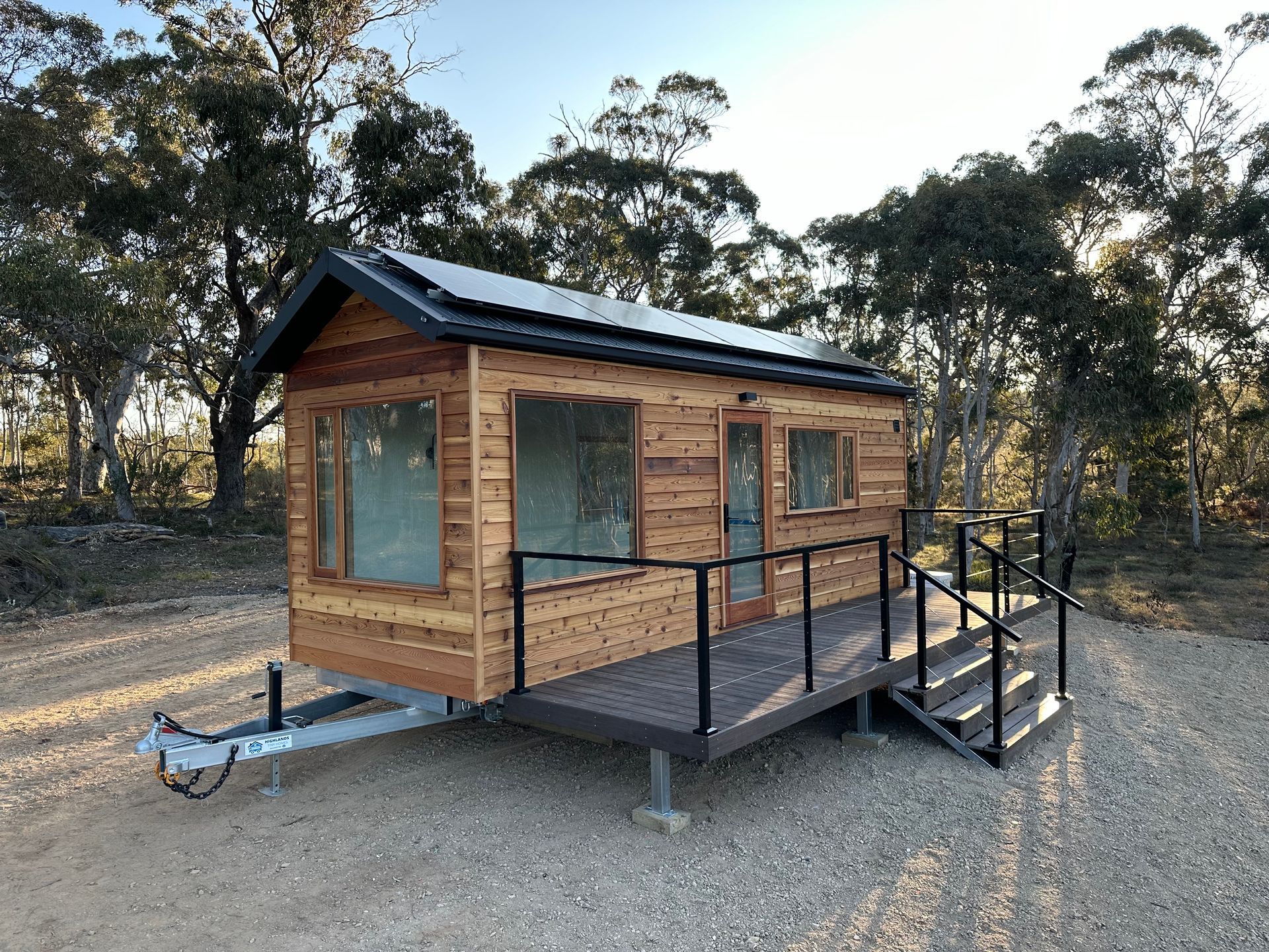 Tiny wooden house on wheels with solar panels, porch, and stairs, parked outdoors.
