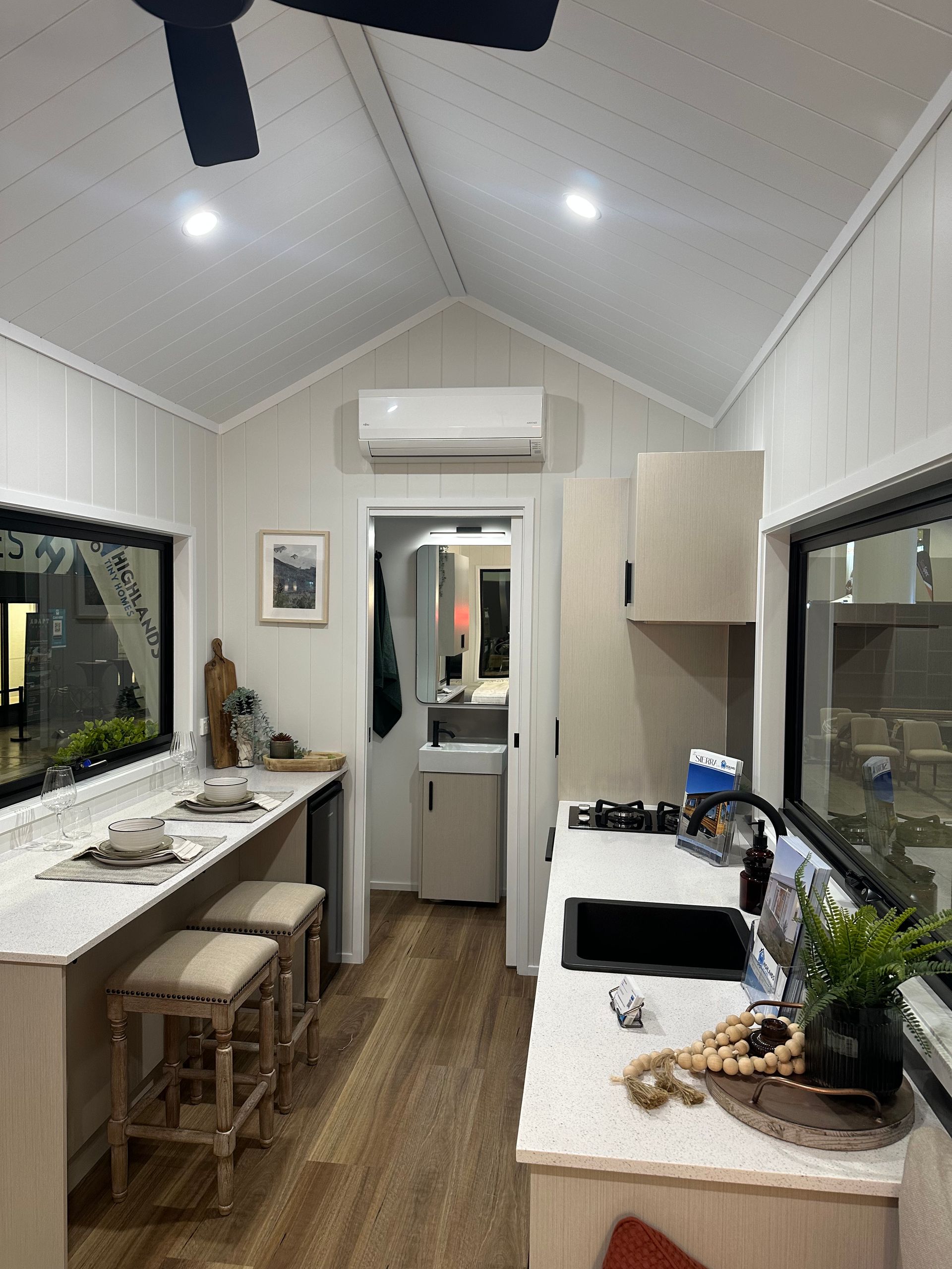 Interior of tiny house, wood floors, white walls, light wood ceiling, ladder to loft, door to bathroom.