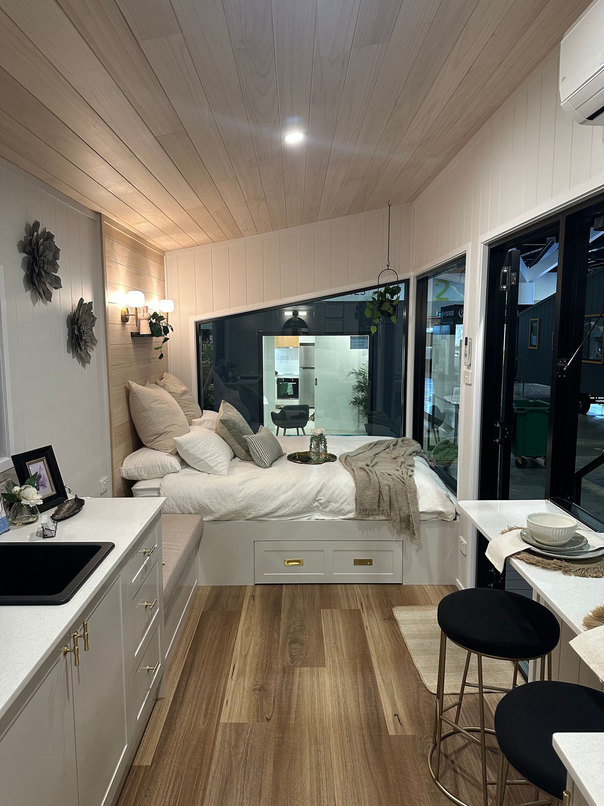 Cozy tiny home interior with a bed by large windows, white cabinets, and light wood flooring.