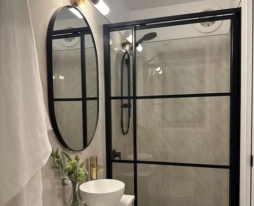 Small modern bathroom with black framed shower, oval mirror, white sink, and light fixture.