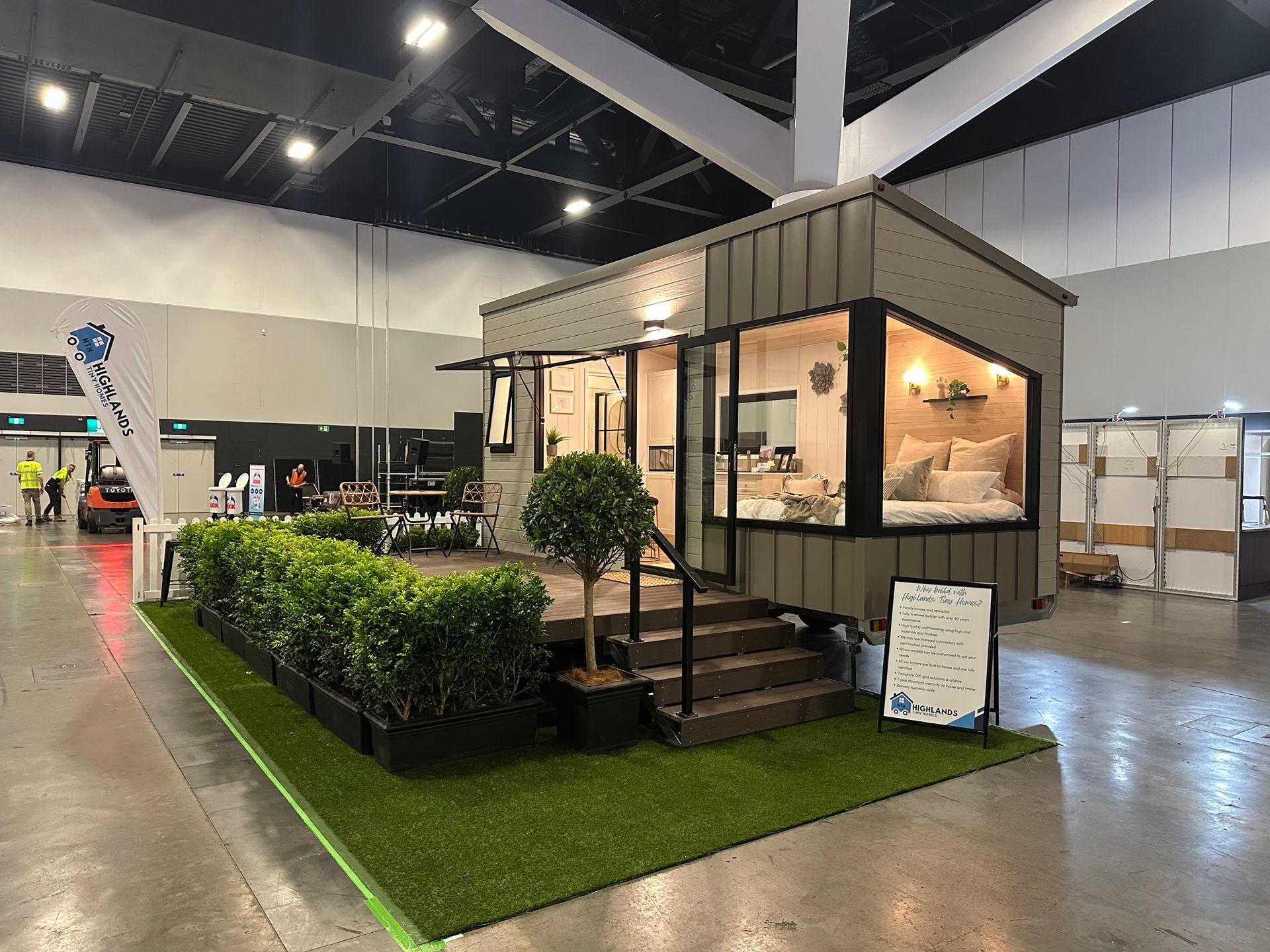 A small modular home at a trade show, with a bed visible inside and a grassy yard display.