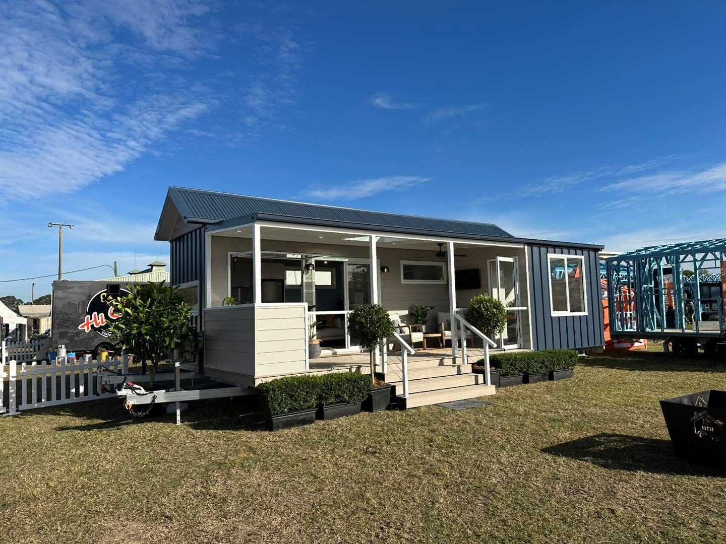 Tiny blue house on trailer, porch with steps, potted plants in front, on grassy area, blue sky.