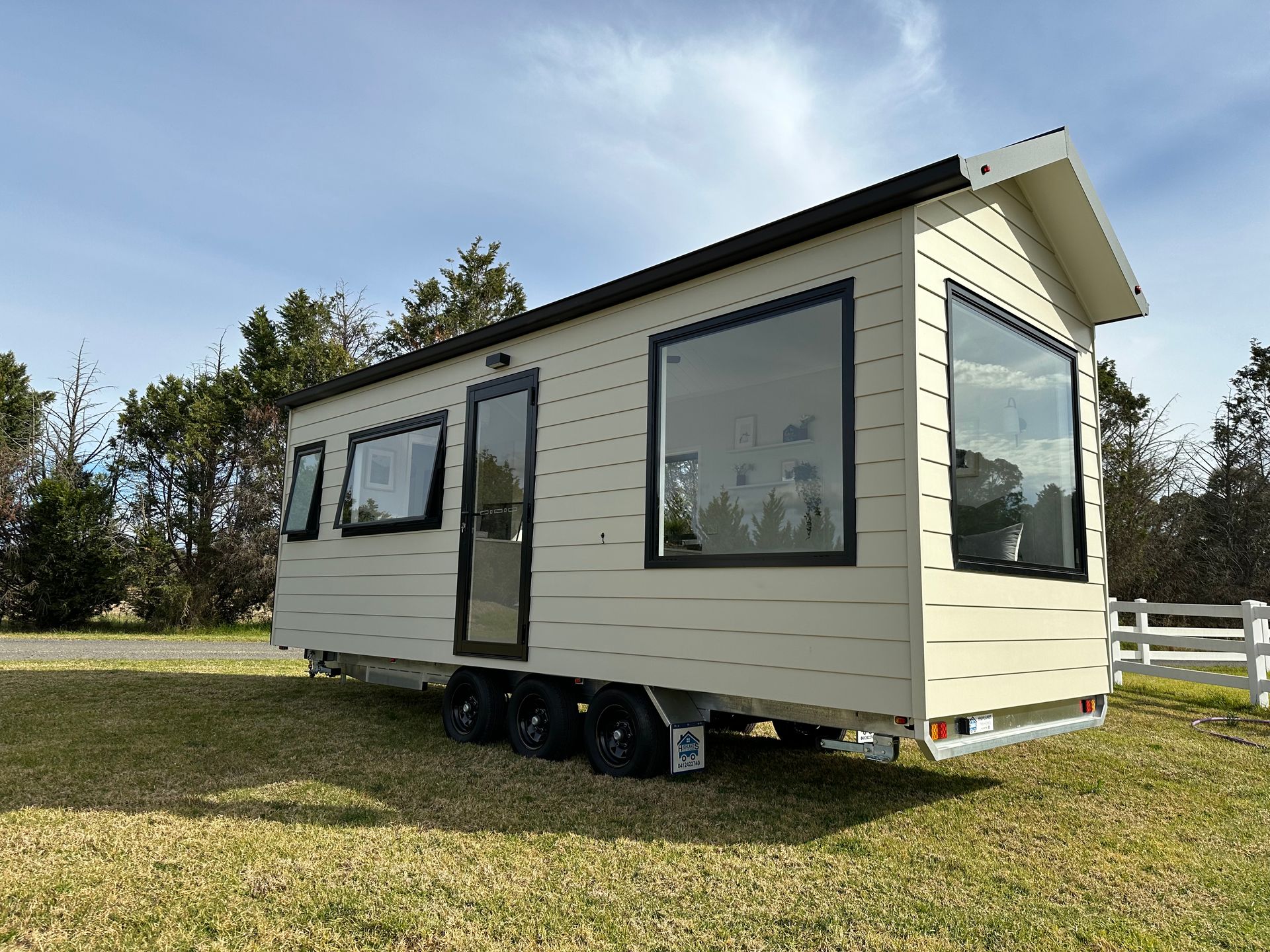 Beige tiny house on a trailer with dark window frames, parked on grass.
