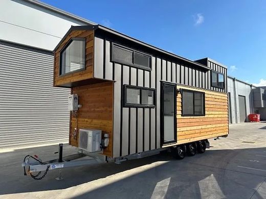 Tiny house on wheels with gray siding and wood accents, parked outside on a sunny day.