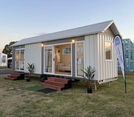 Tiny house, light gray exterior, sliding glass doors, set in a grassy field, steps, with a banner.