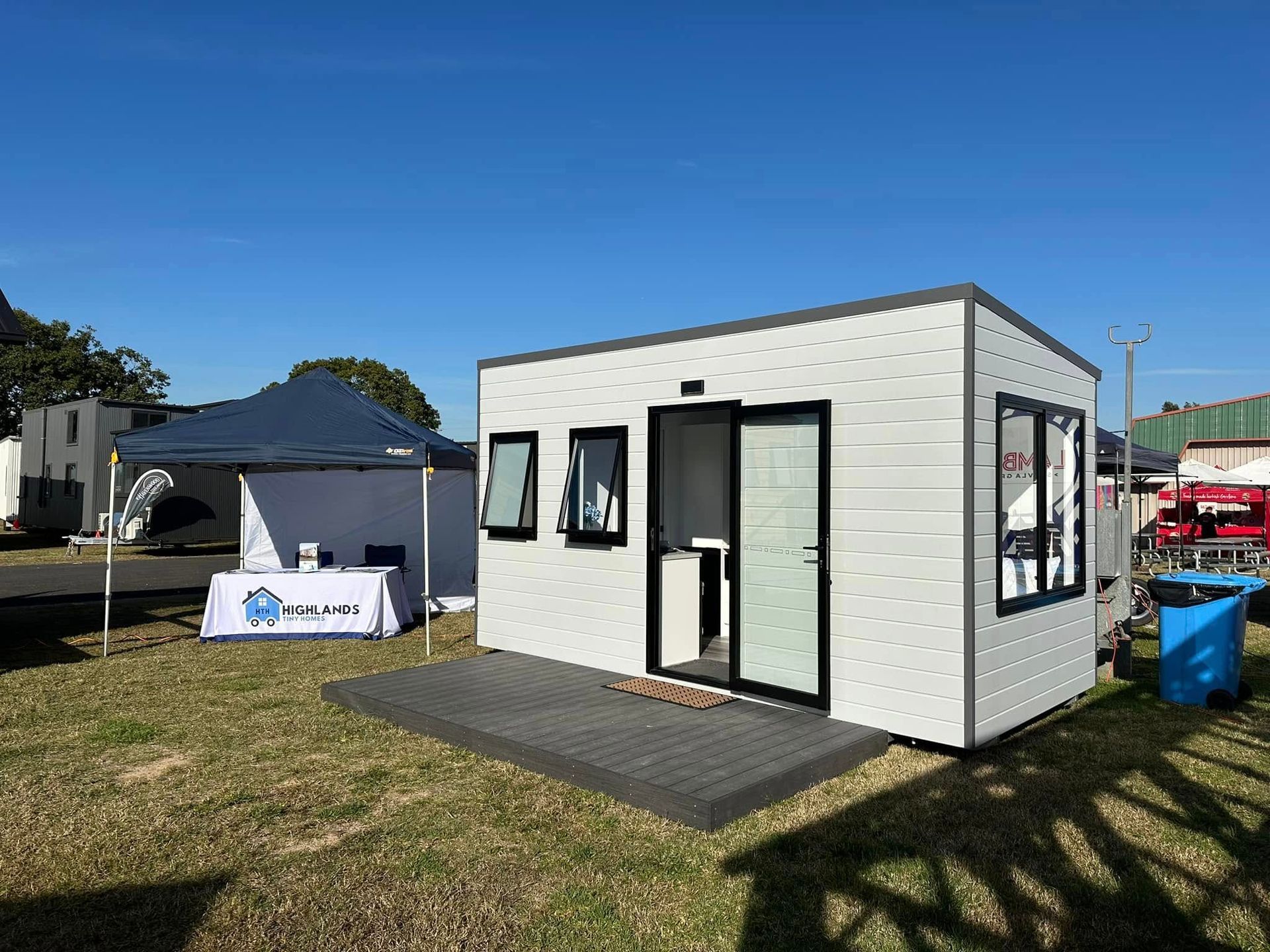 White modular building with black trim, door open. Display set up on a grassy field, under blue sky.