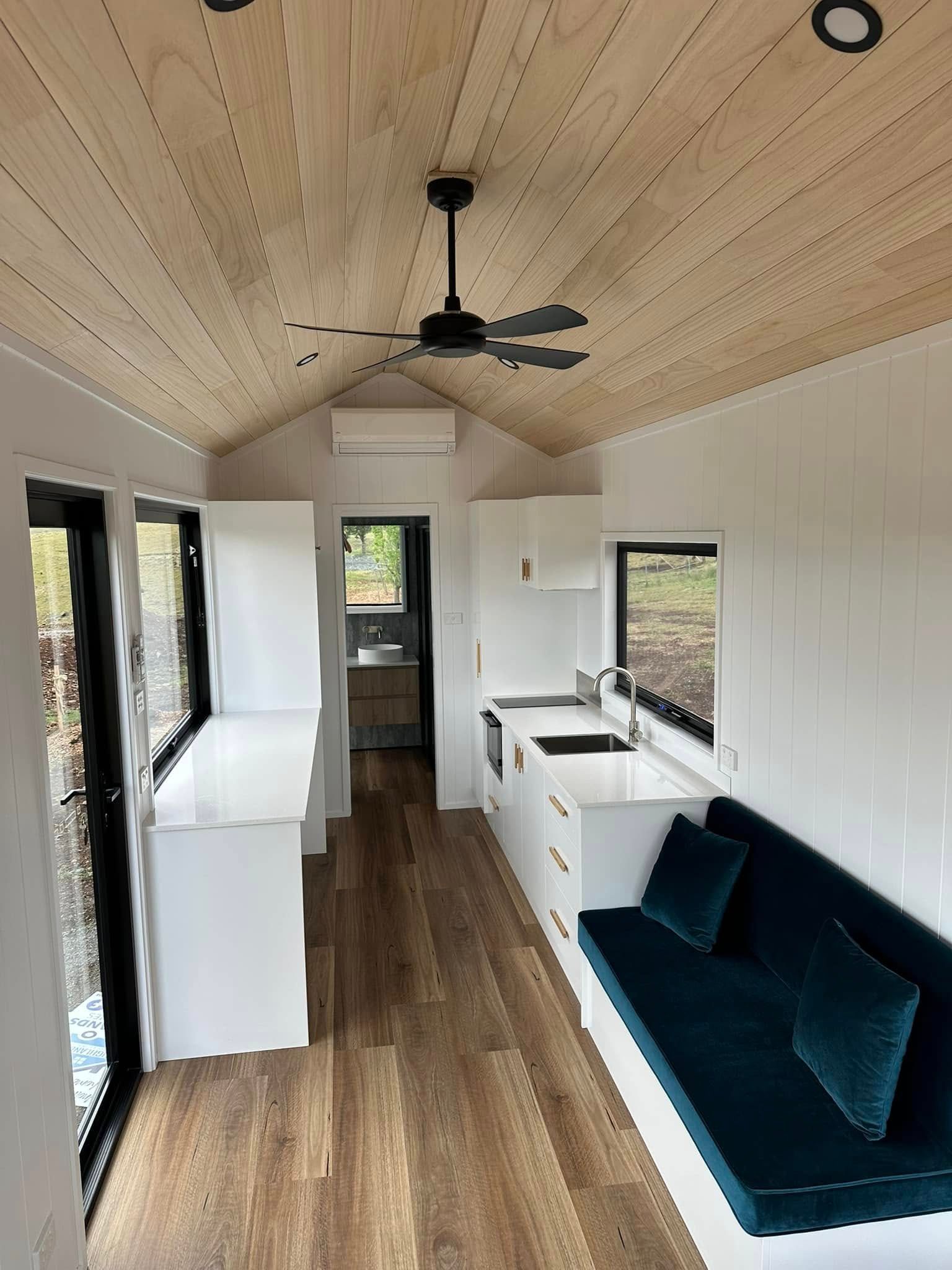 Interior of tiny home with white walls, wood ceiling, dark blue couch, kitchen, and bathroom.
