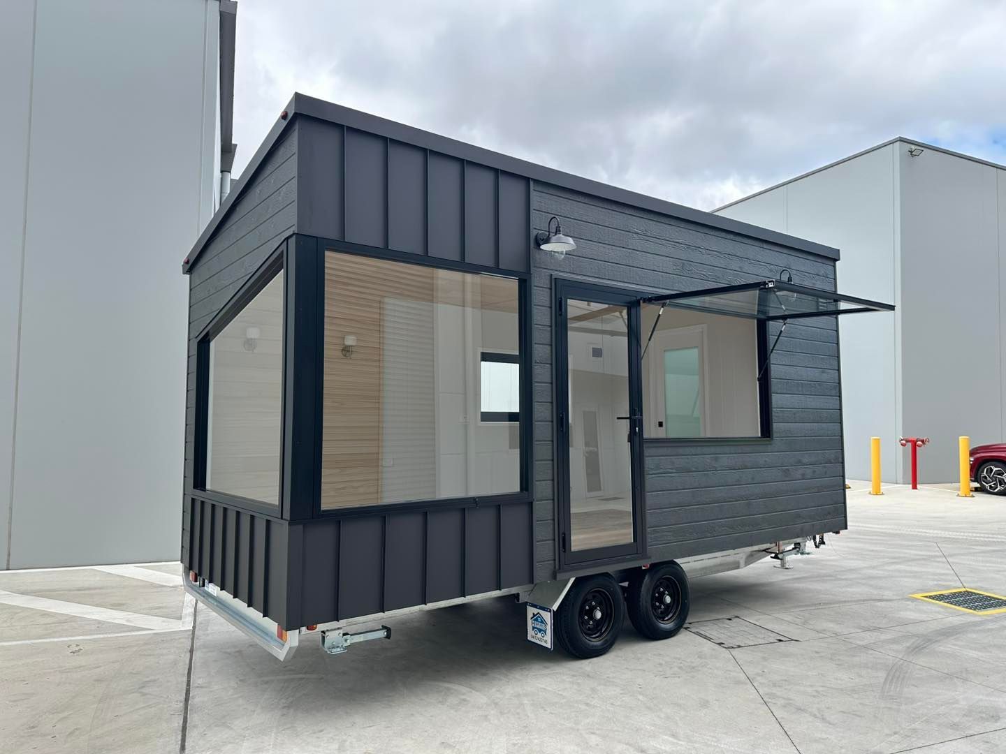 Tiny wooden house on wheels with black window and trailer hitch on a grassy field.