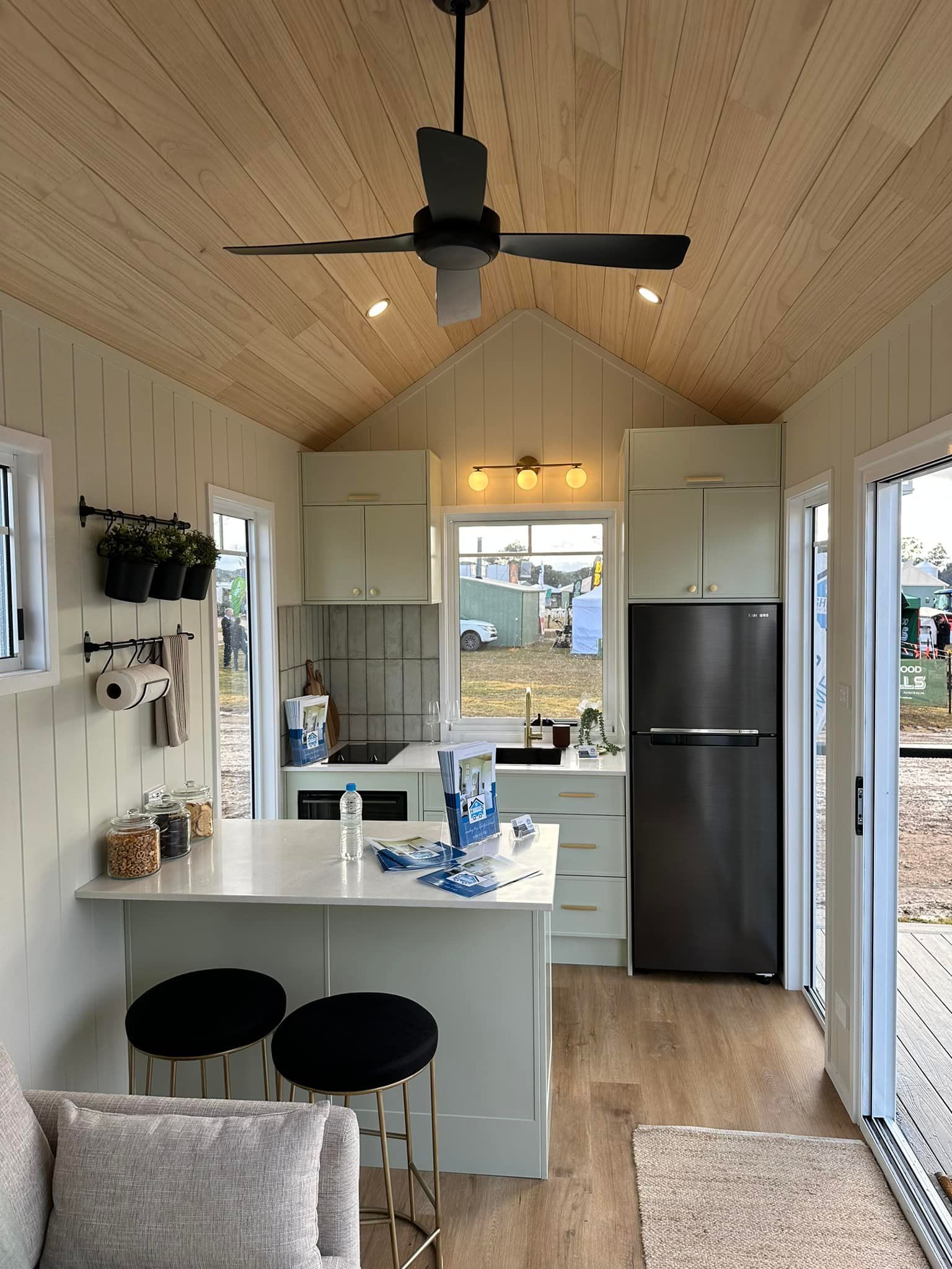 Interior view of a tiny house kitchen. White cabinets, island with black stools, refrigerator, and a wooden ceiling.
