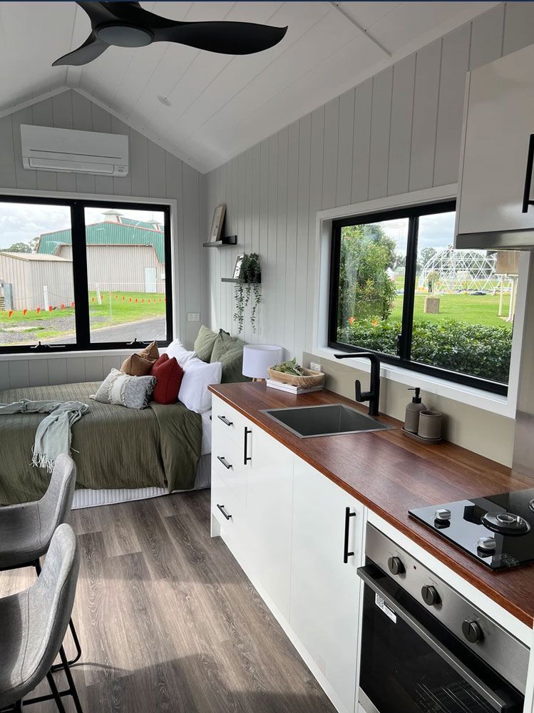 Small, bright tiny home interior with kitchen, bed, and large windows.
