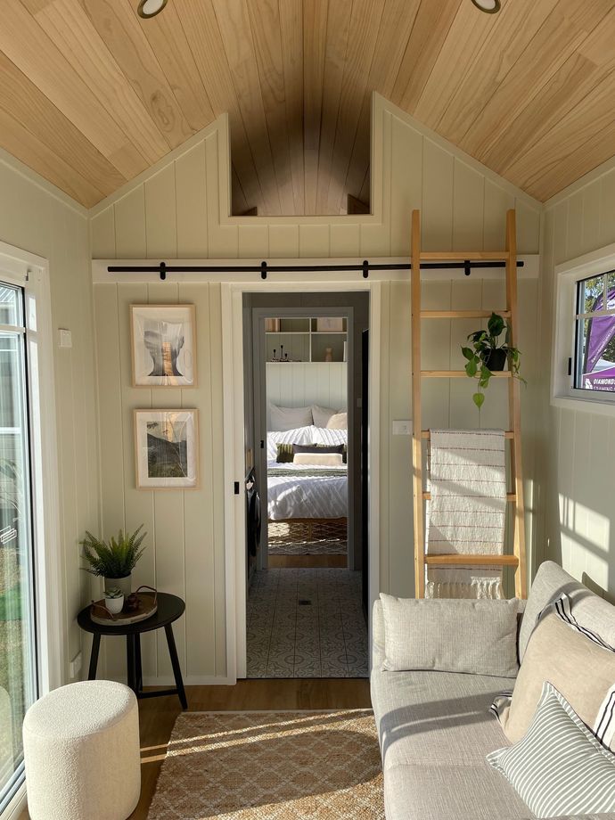 Cozy interior of a tiny home with a couch, ladder, artwork, and a view into a bedroom.
