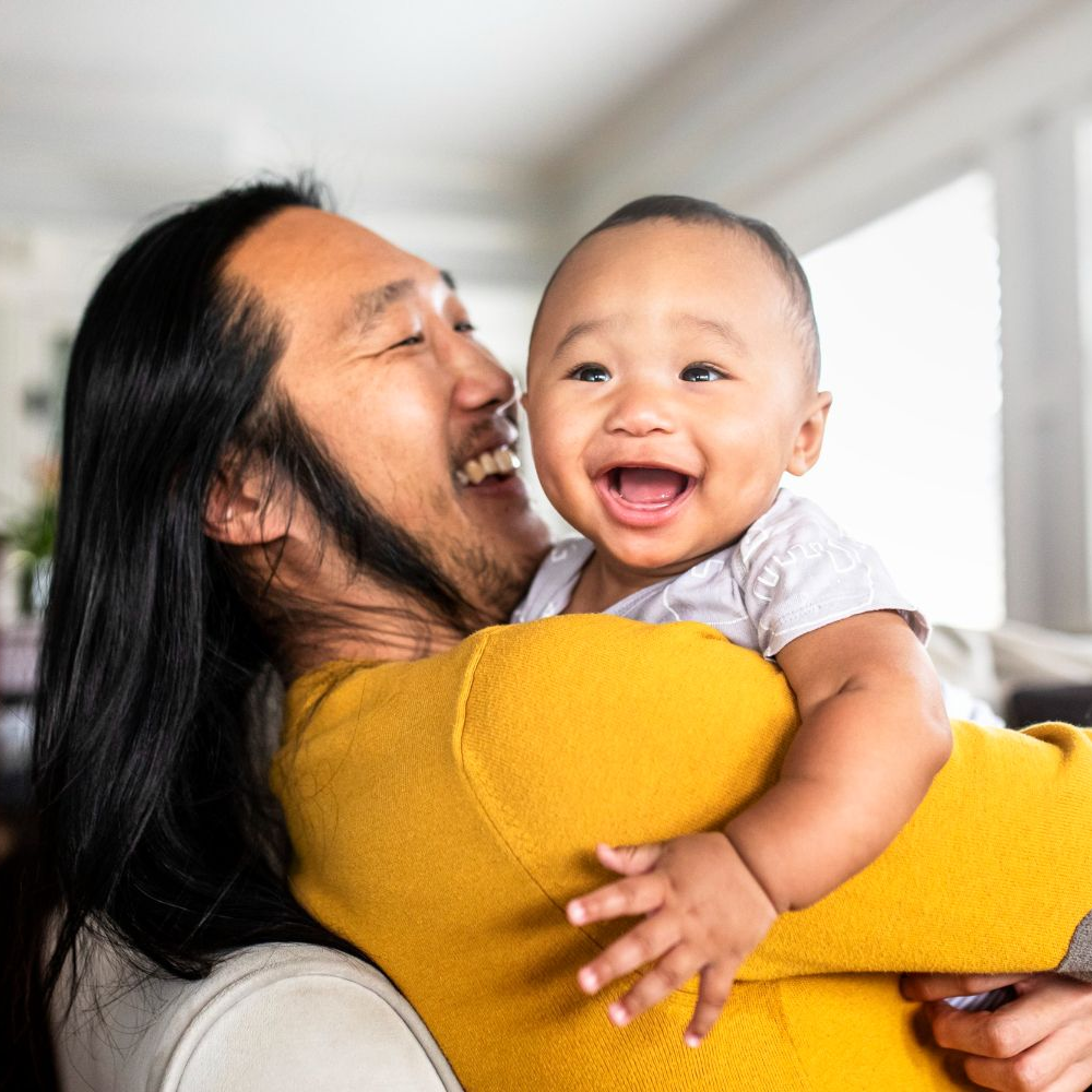 Two people smiling and hugging indoors, with a baby in a yellow sweater laughing in the foreground