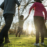 Three people holding hands and running through a sunlit park with trees and grass