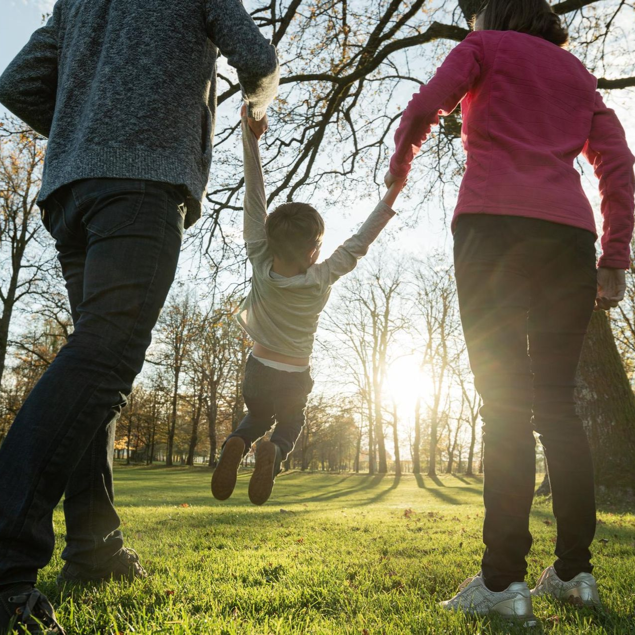 Two adults swinging a child by the hands in a sunny park at sunset