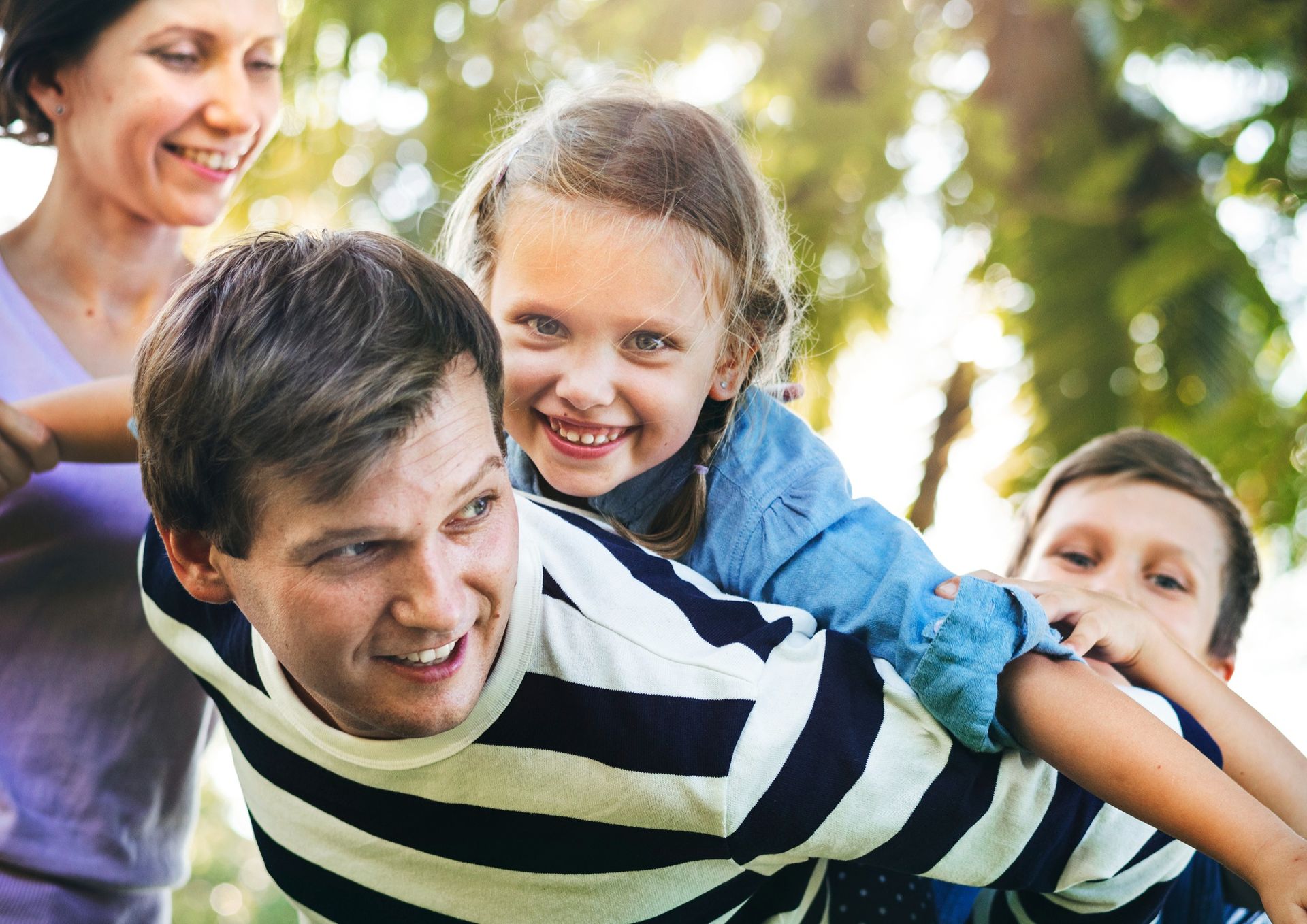 Family playing together outdoors, with a smiling child riding on a man’s back