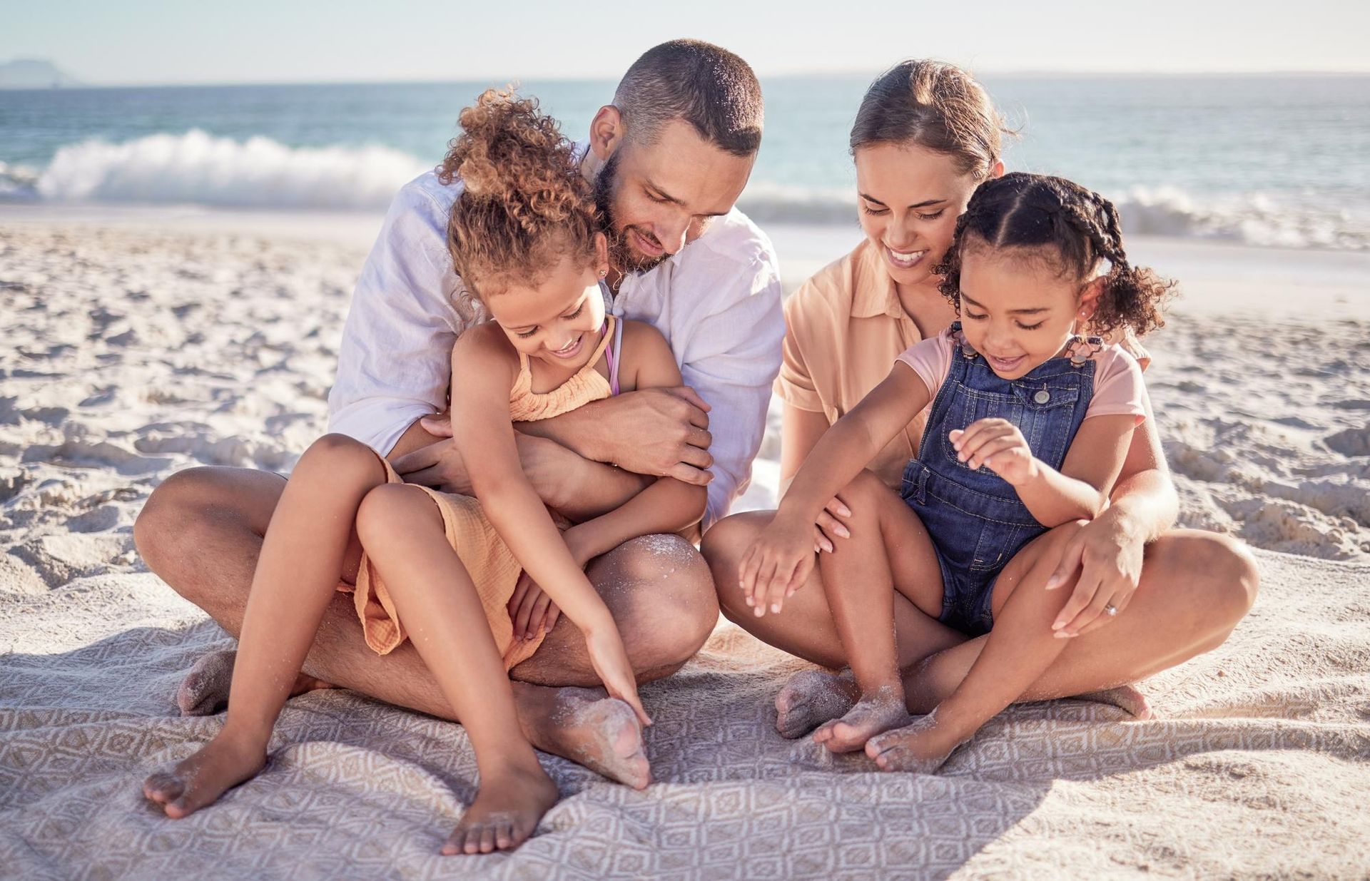 Family sitting on a sandy beach, smiling and holding two children near the ocean